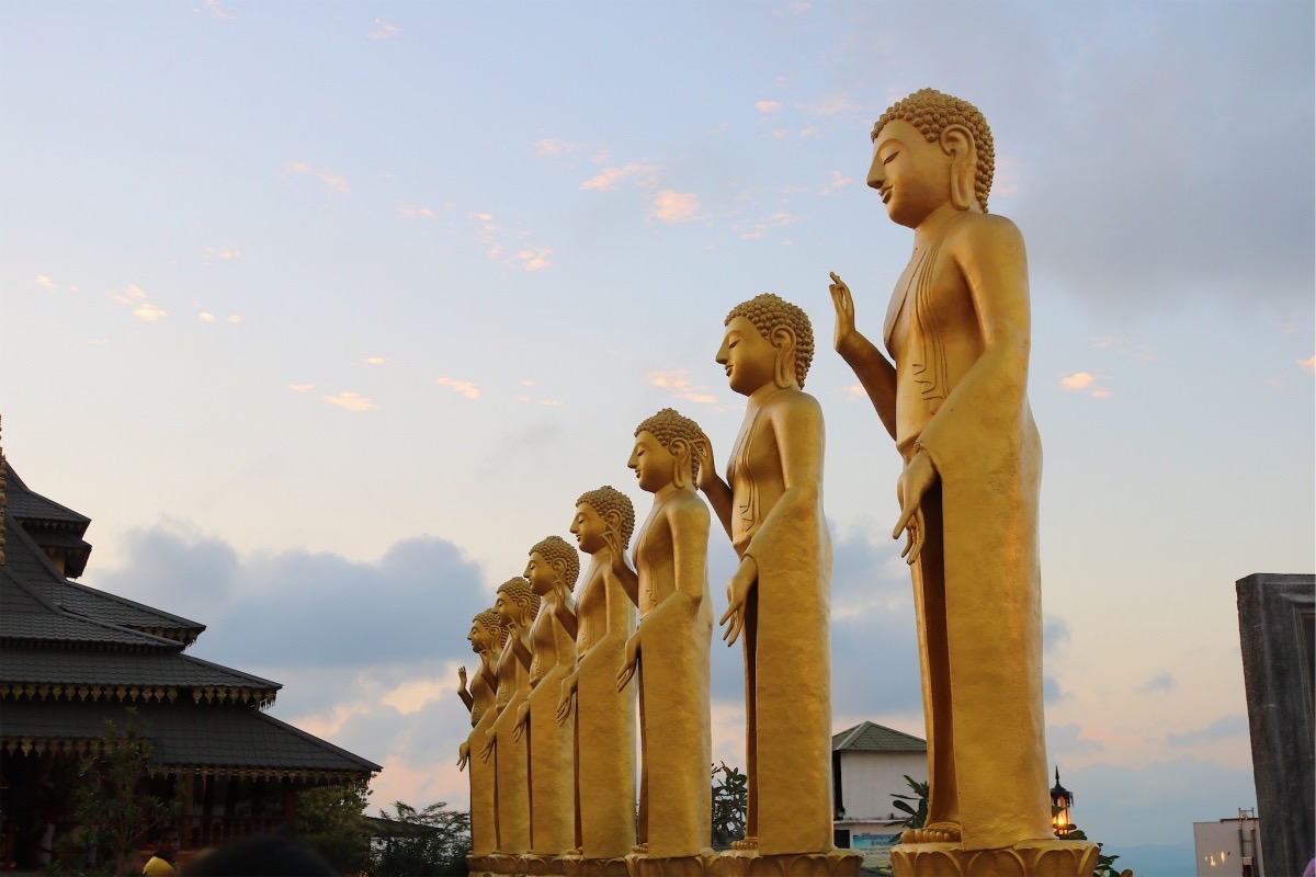 Seven golden Buddha statues standing in a row outdoors against a sky with clouds, with traditional buildings nearby.