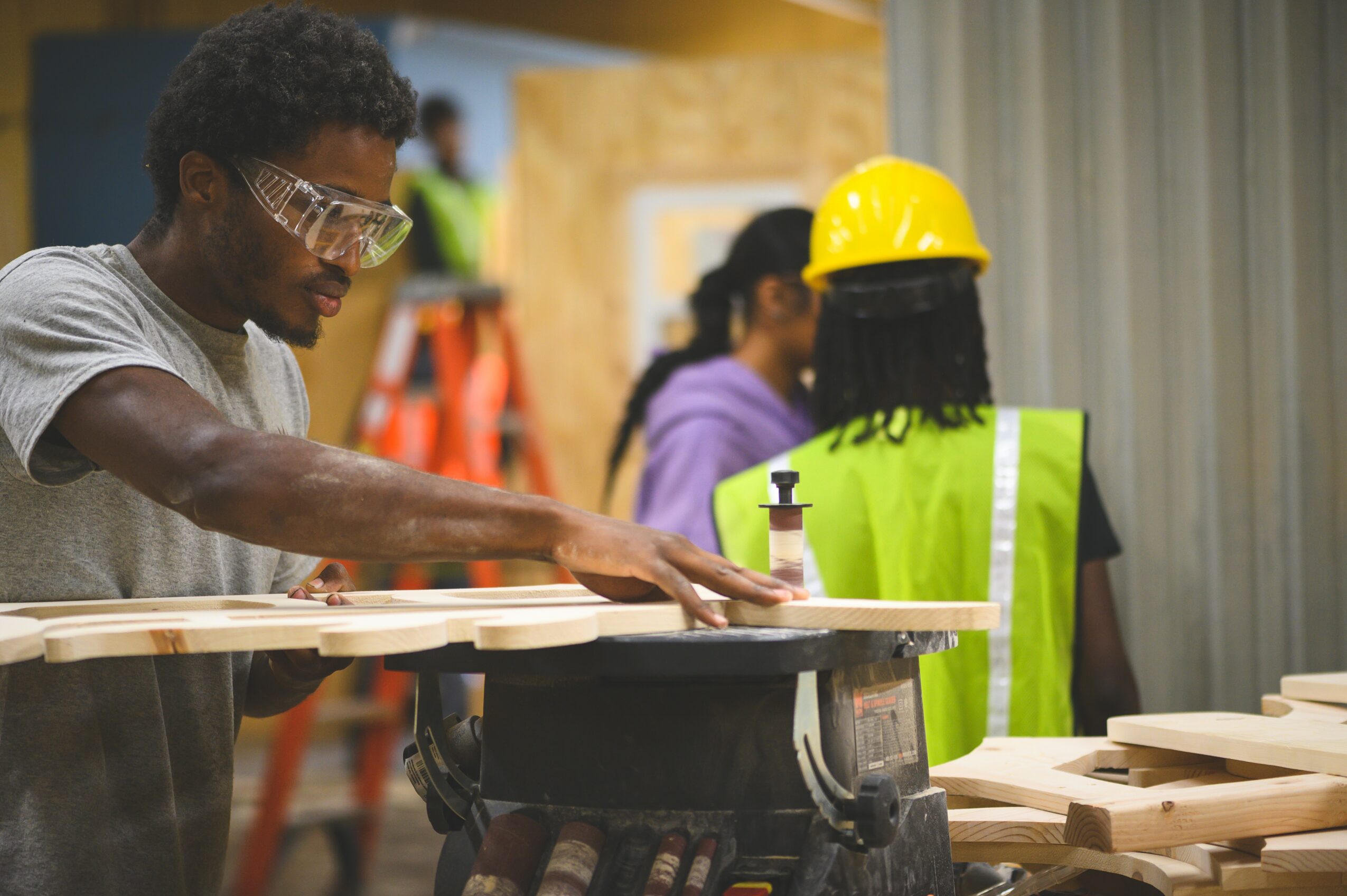 Person working with wood at a construction site, wearing safety glasses and a gray shirt.