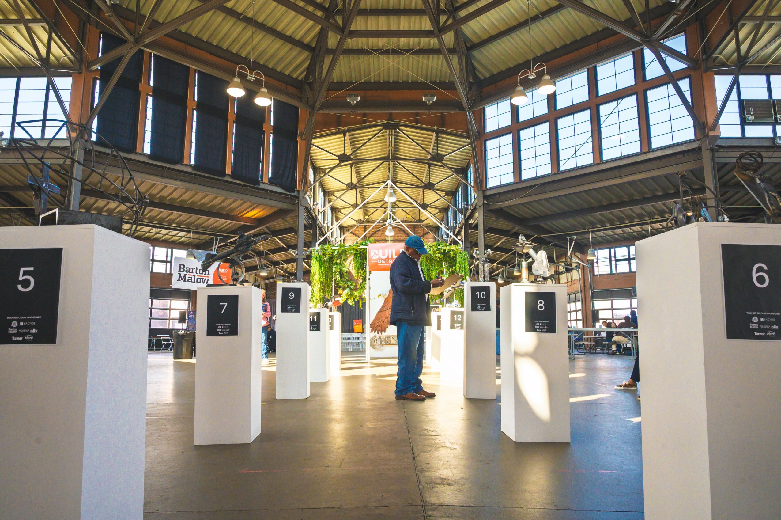 Person standing in an exhibition space with numbered display stands and hanging plants, inside a large glass and wood structure.