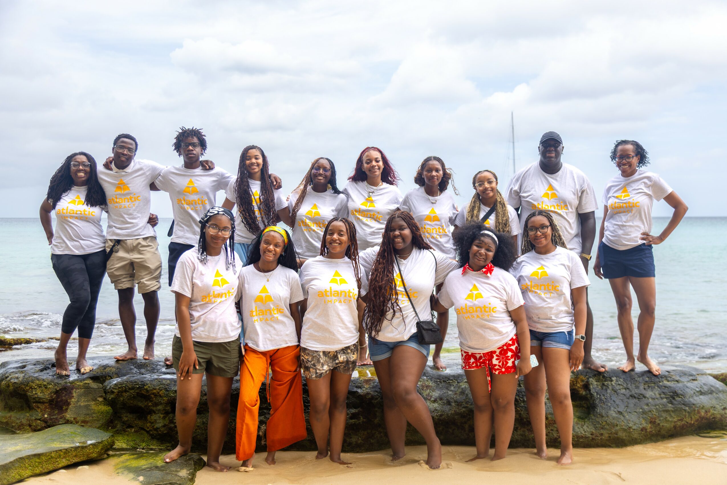 Group of 14 people standing on rocks at the beach, some with hands on hips, smiling, with ocean and cloudy sky in background.