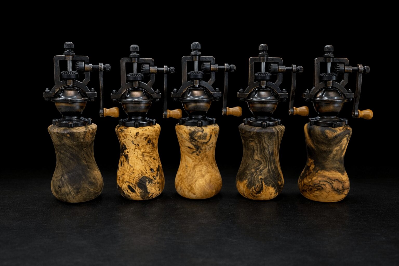 Five vintage coffee grinders with wooden bases and metal handles, lined up on a black surface against a black background.