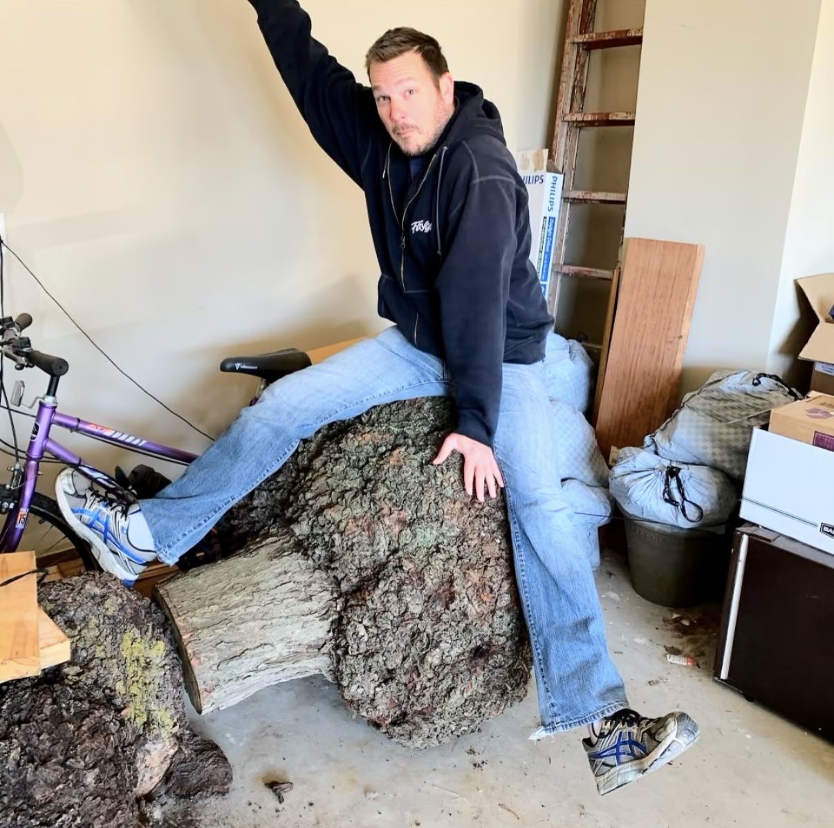 Person sitting on a large fallen tree trunk indoors, with one arm raised and legs spread apart.