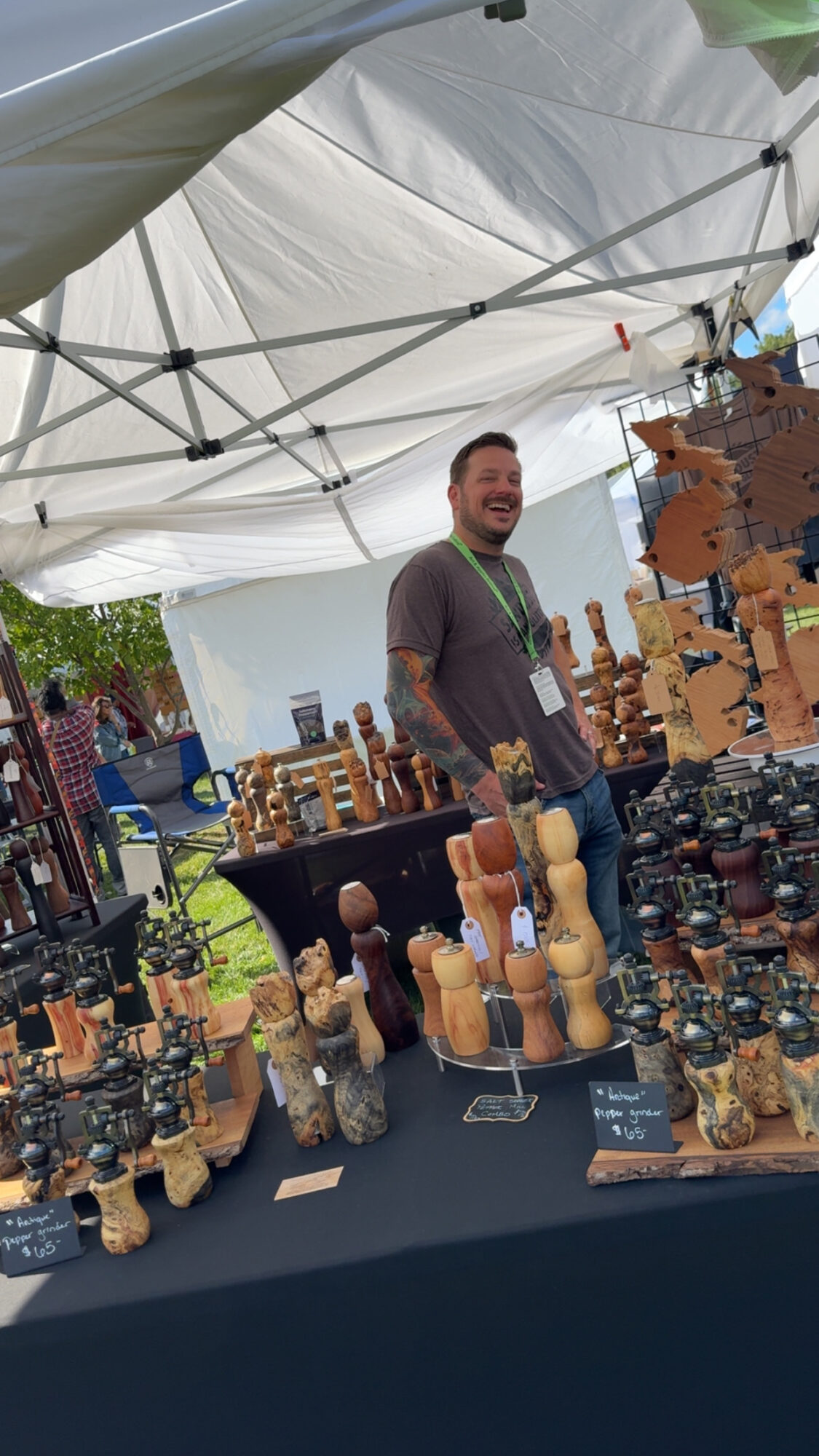 Man standing behind a table with wooden and ceramic items at an outdoor market stall under a white canopy.