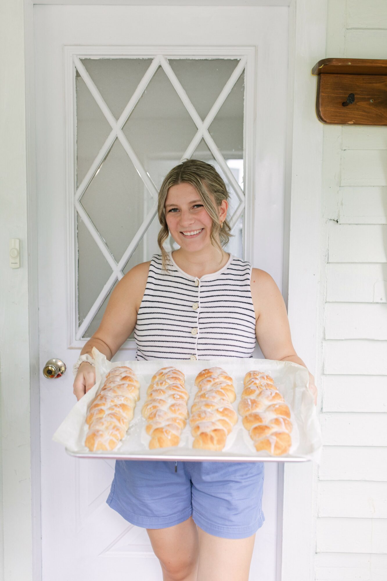 Young woman holding a tray of pastries, smiling, standing in front of a door with a diamond pattern, wearing a striped top and blue shorts.