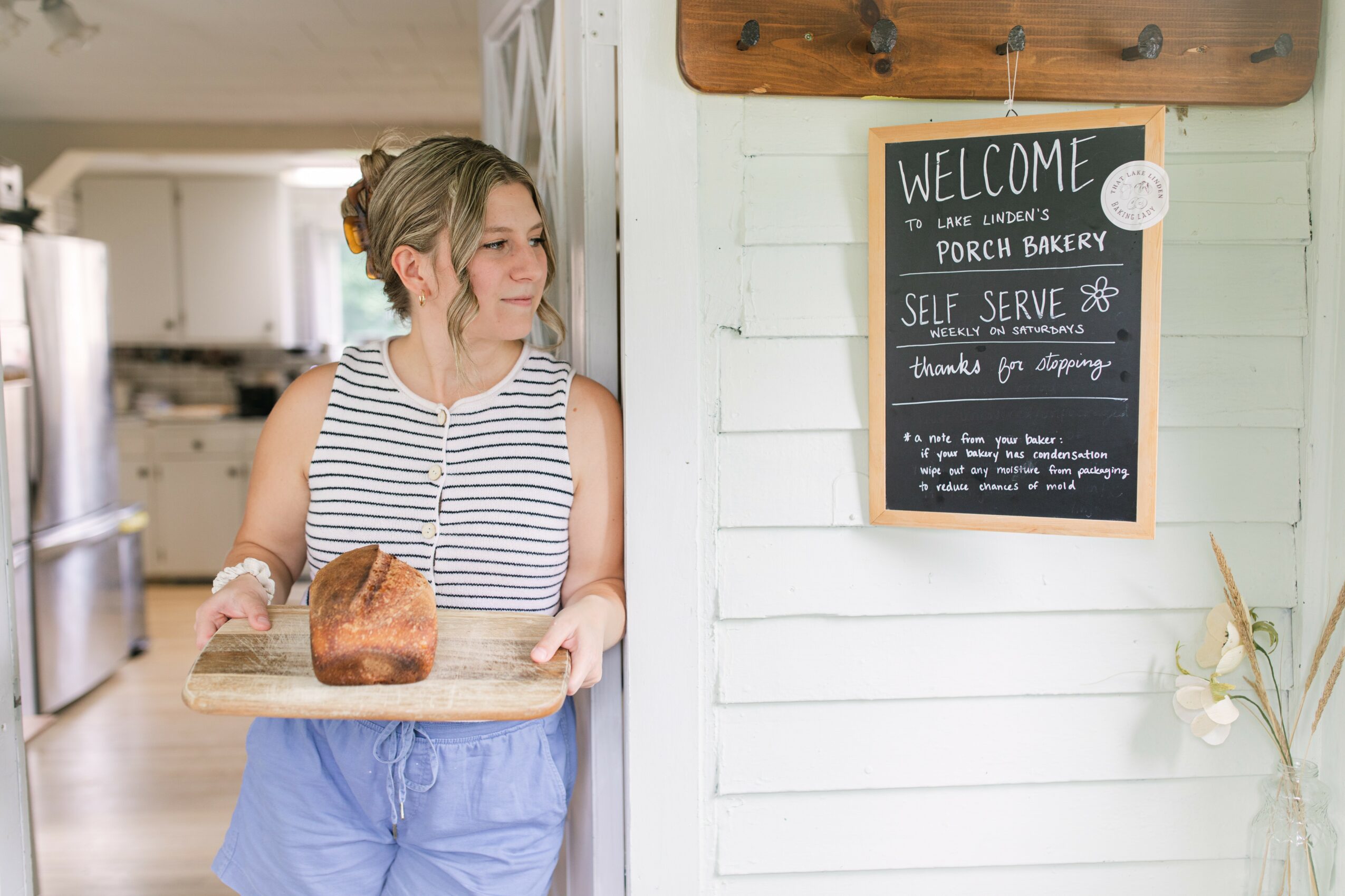 Young woman holding a loaf of bread on a tray inside a bakery, standing near a white wall with a welcome sign.