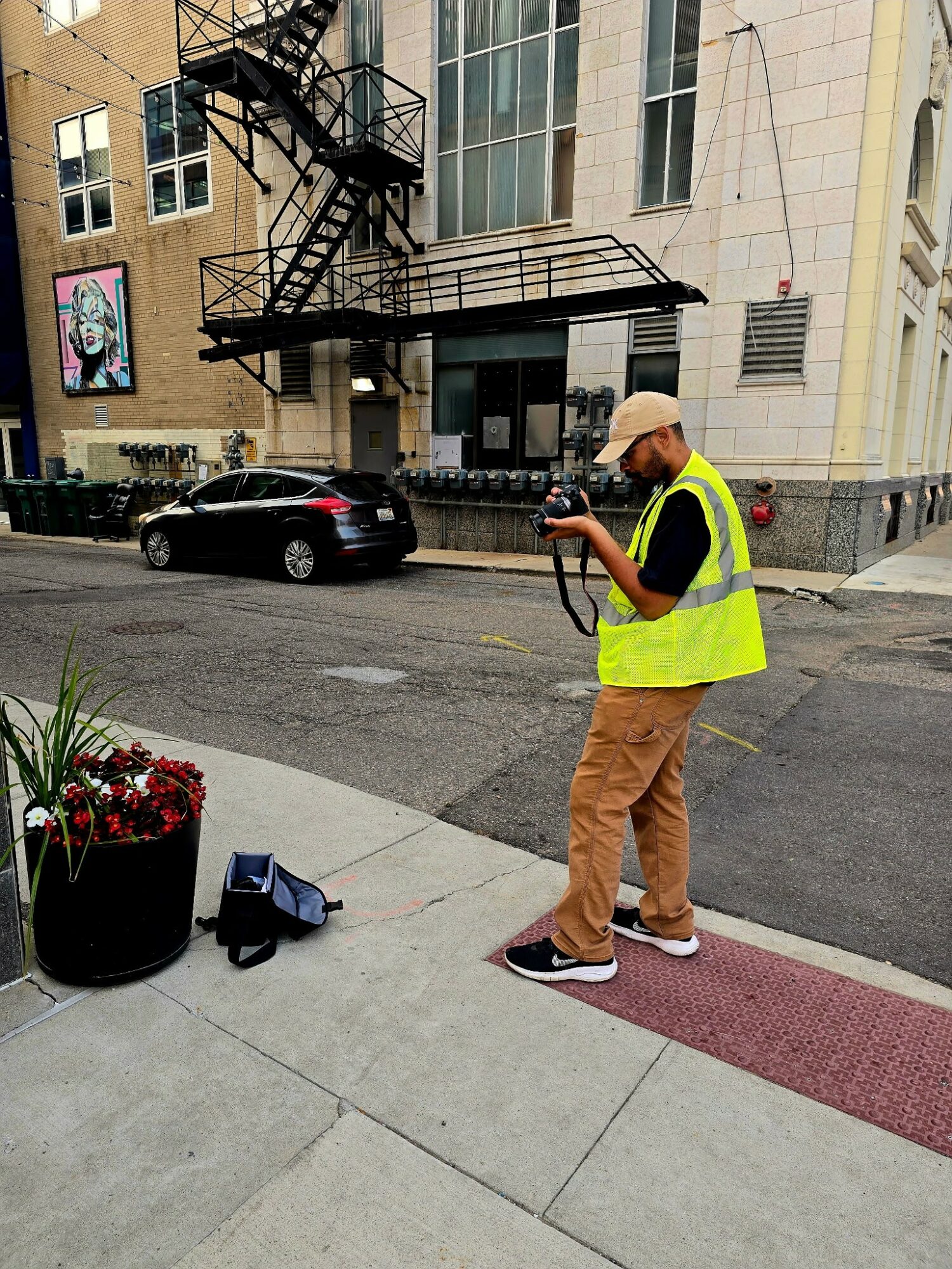 Person in yellow vest and beige pants standing on sidewalk, looking at phone, with potted flowers nearby, urban street background.