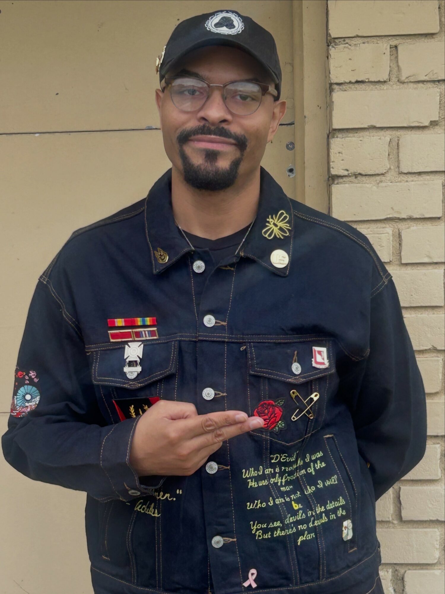 Man wearing glasses, a cap, and a dark jacket with patches and writing, standing outdoors against a brick wall.
