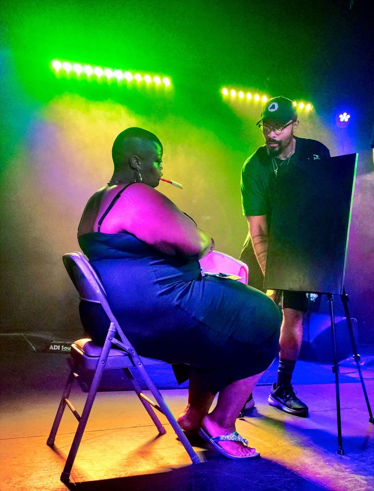 Two people on stage with colorful lighting, one sitting on a chair and the other standing, near an easel or board.