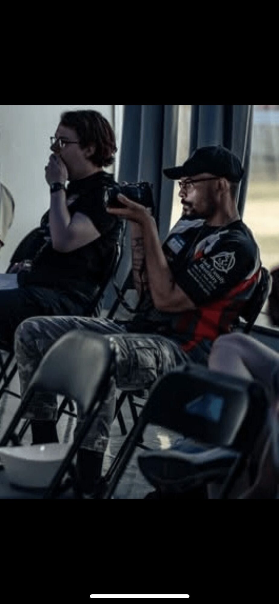 Two men sitting indoors, one taking a selfie with a smartphone, the other wearing glasses and a cap, with curtains in the background.