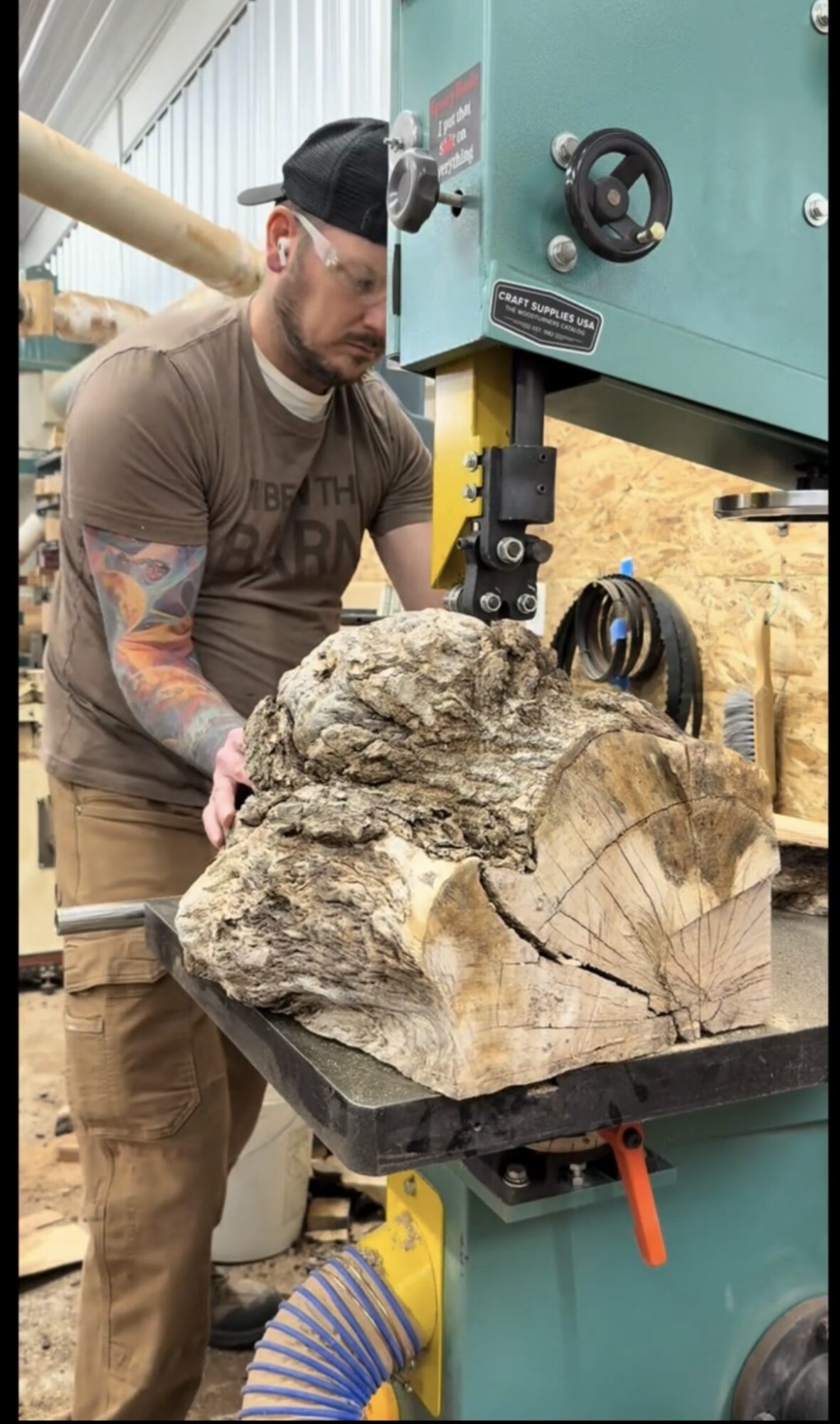 Man with tattoos operating a large machine to cut a big log of wood, in a workshop setting.