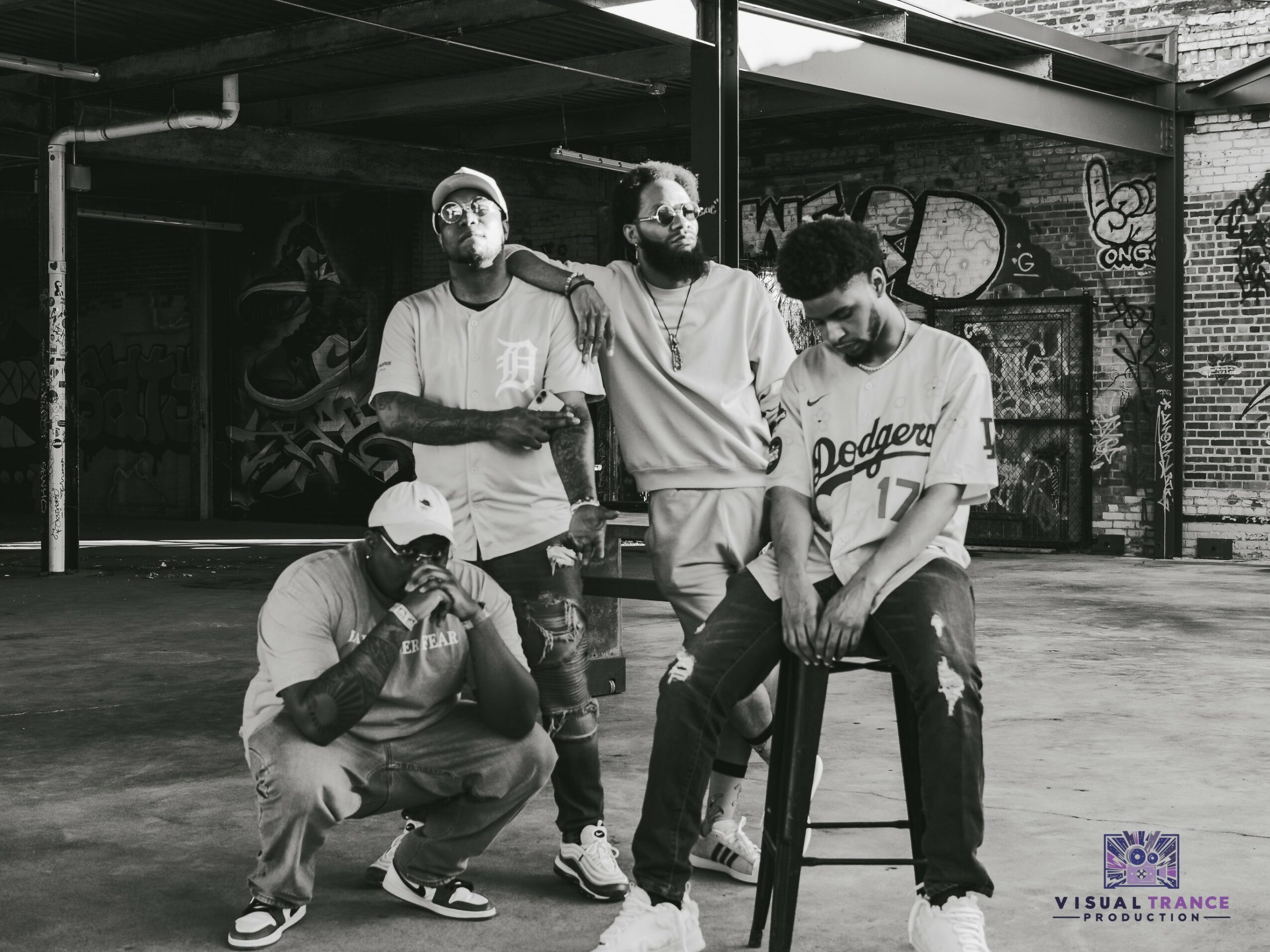 Four young men pose outdoors in front of a graffiti-covered wall, with one sitting on a stool and others standing nearby.