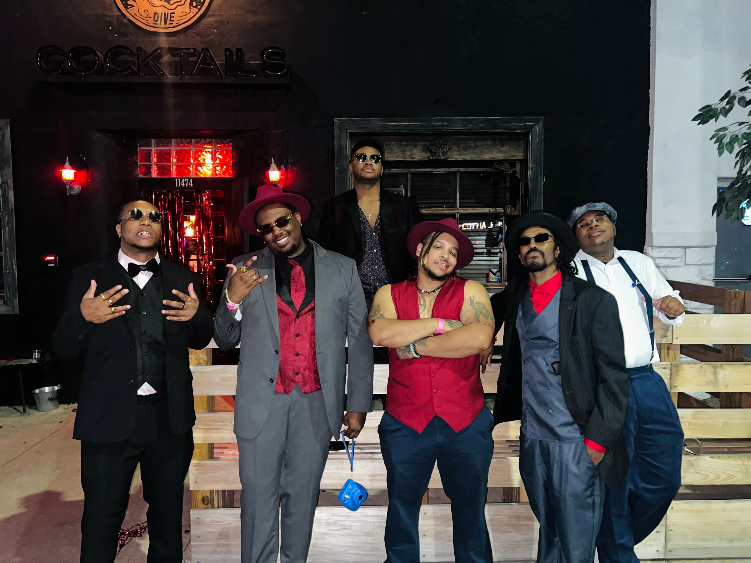 Group of six people standing outside a building, some wearing sunglasses and colorful clothing, posing for a photo.