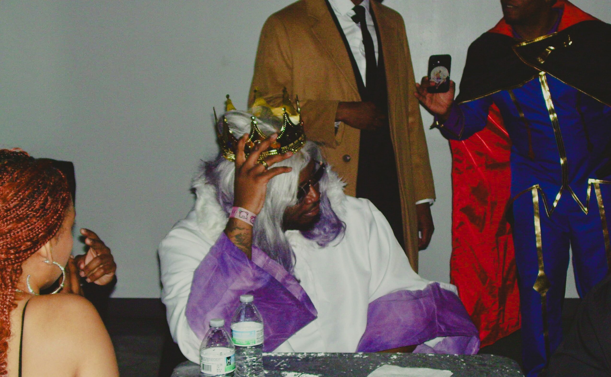 Person with long gray hair and crown, wearing white and purple, seated at a table with water bottles, surrounded by others.