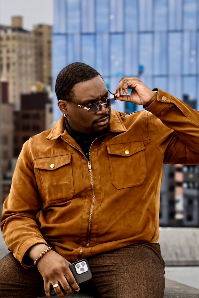 Person adjusting sunglasses on city rooftop with tall buildings and glass skyscraper in background.