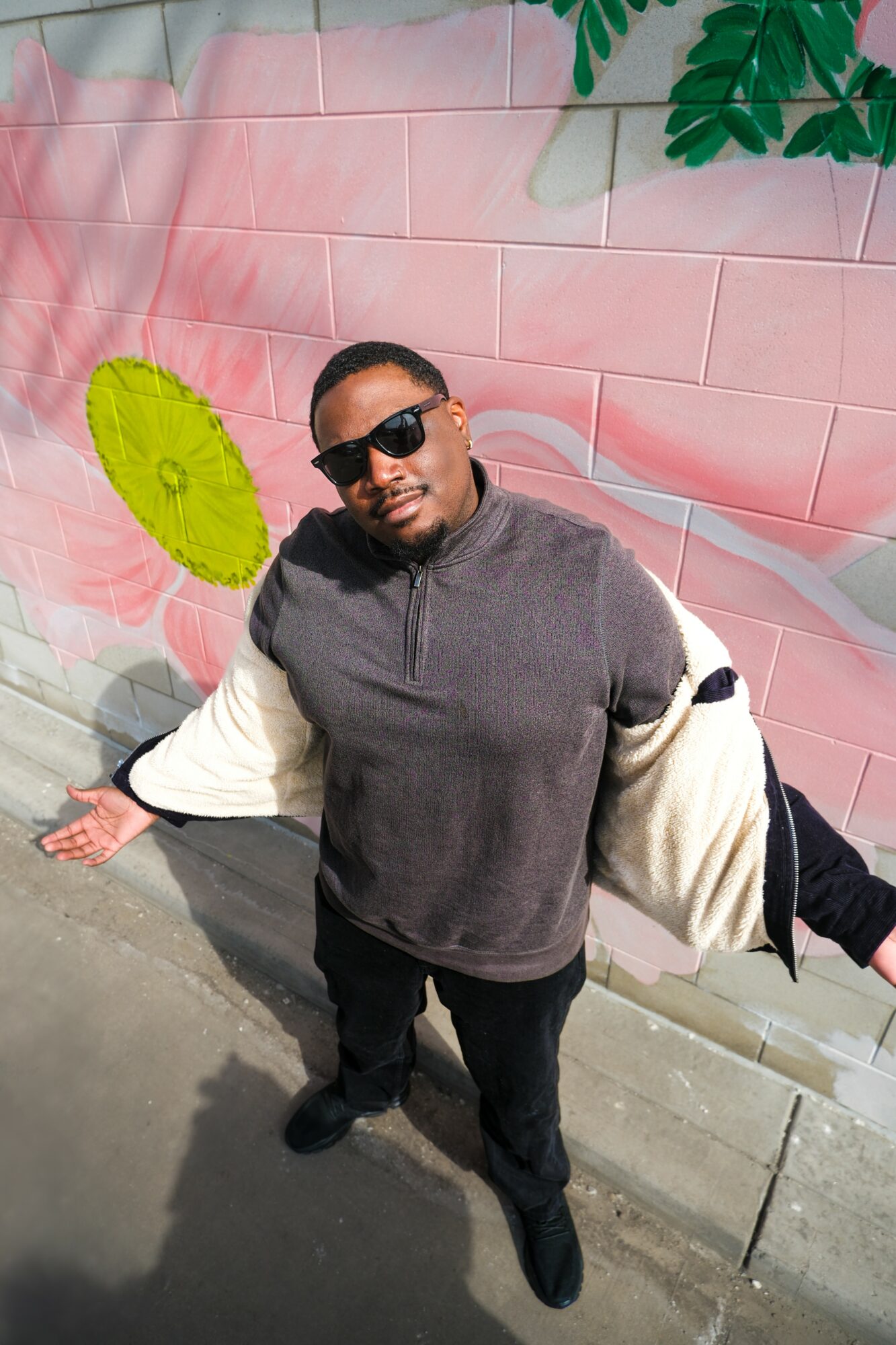 Person wearing sunglasses and a gray and white sweatshirt, standing outdoors with a pink mural and green leaves in background.