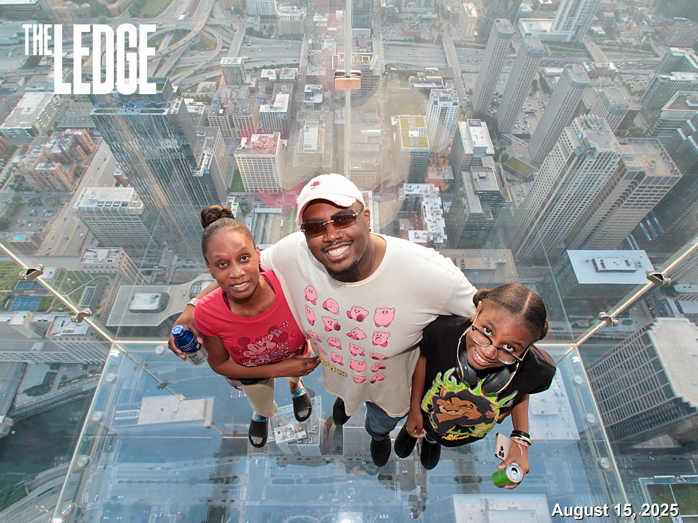 Three people stand on a glass platform high above a city, smiling and looking up at the camera, with tall buildings around.
