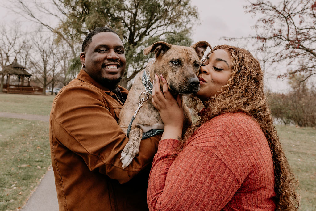 Two people and a dog outdoors in a park, smiling and touching the dog, with trees and grass in the background.