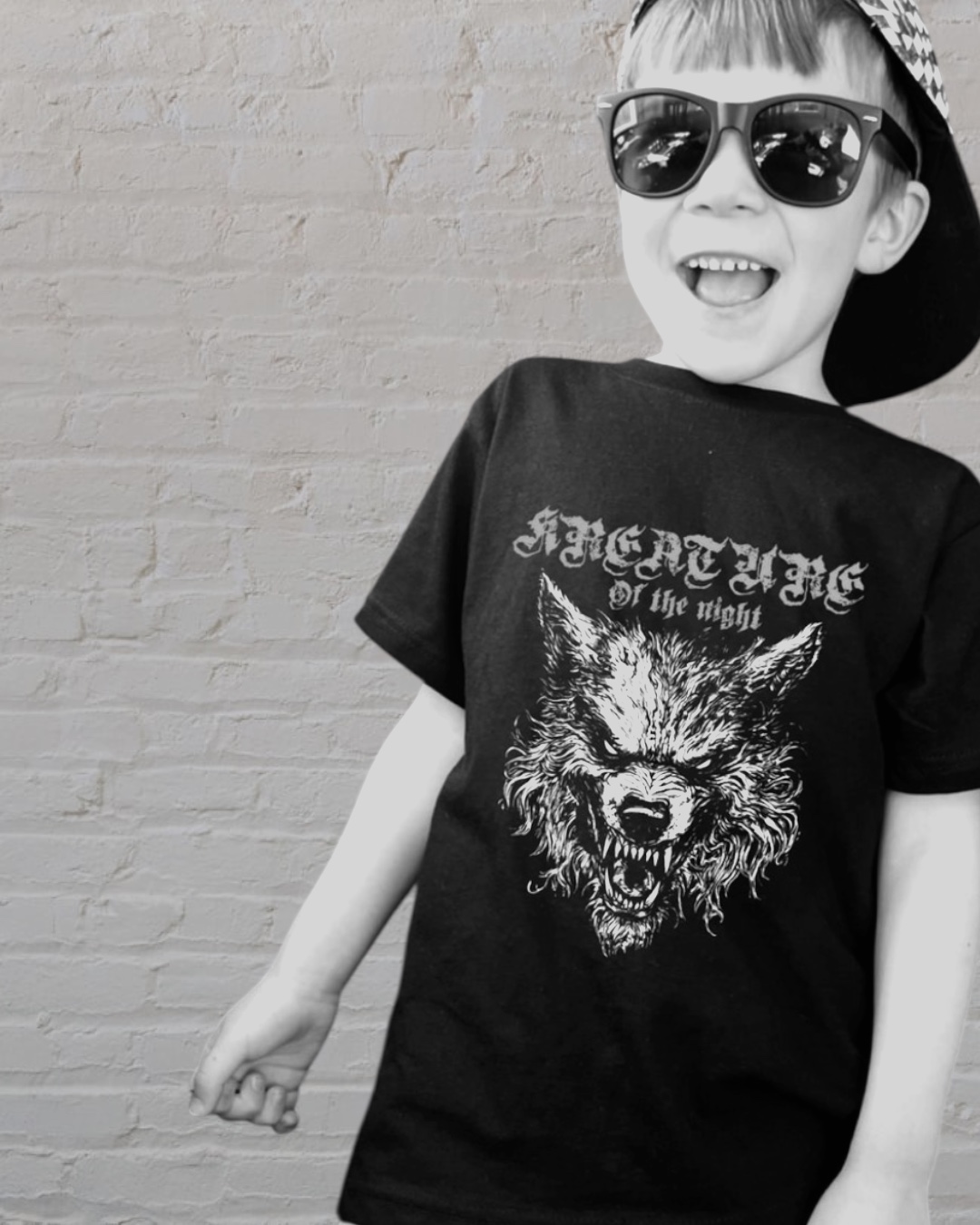 Child wearing sunglasses, a cap, and a graphic T-shirt with a wolf design, smiling and standing against a brick wall.