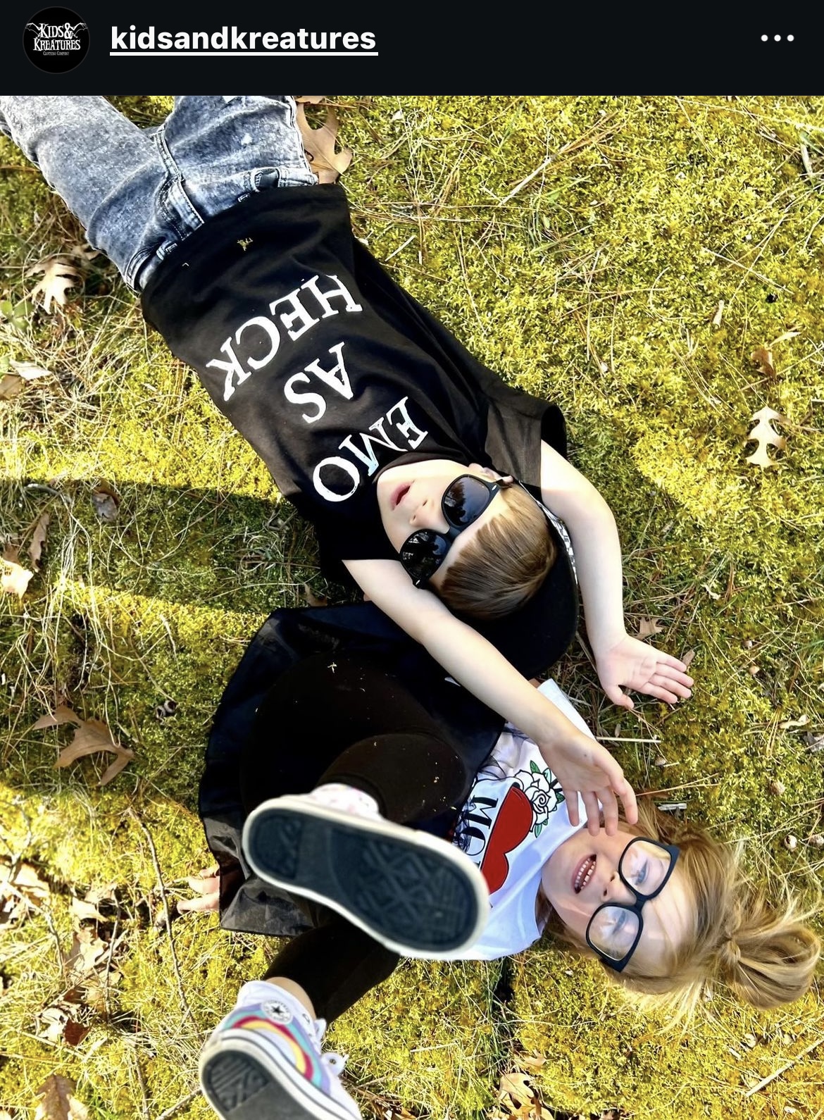 Two children lying on grass, one wearing glasses and a white shirt, the other in a black shirt, outdoors.