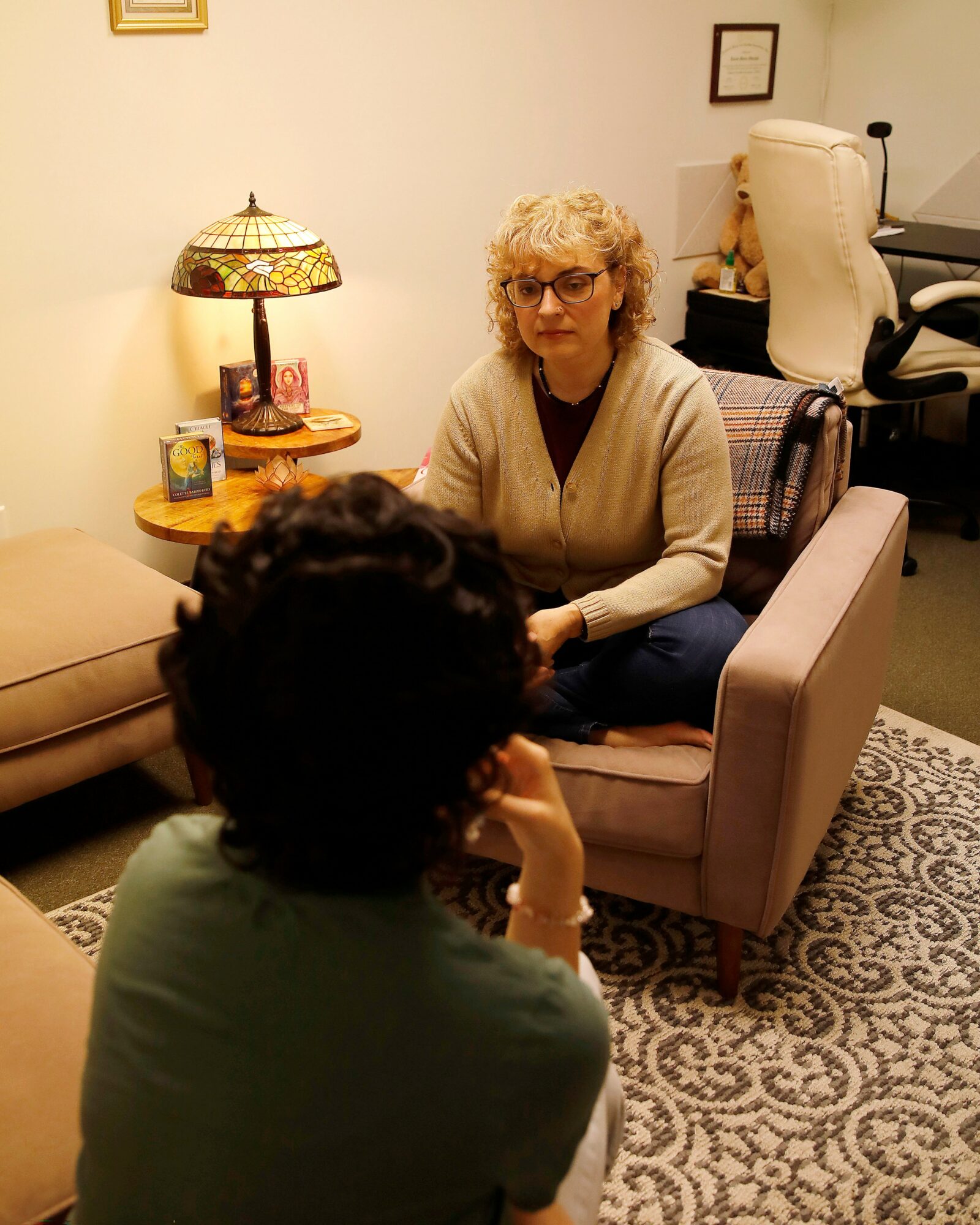 Two women sit and converse in a cozy room with a patterned rug, lamp, and framed pictures on the wall.