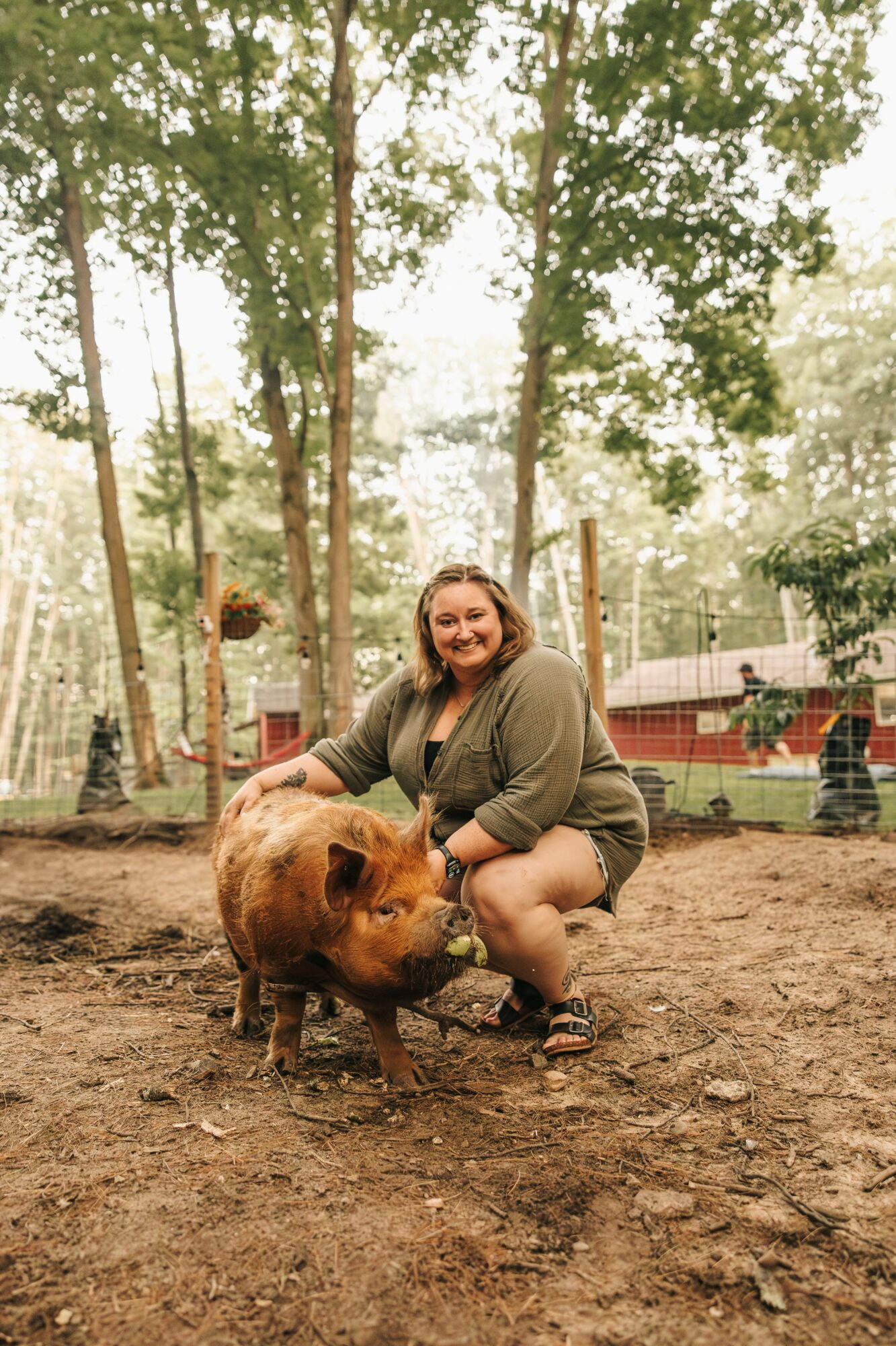 Woman crouching next to a pig in a wooded outdoor area, trees and a fence in the background.