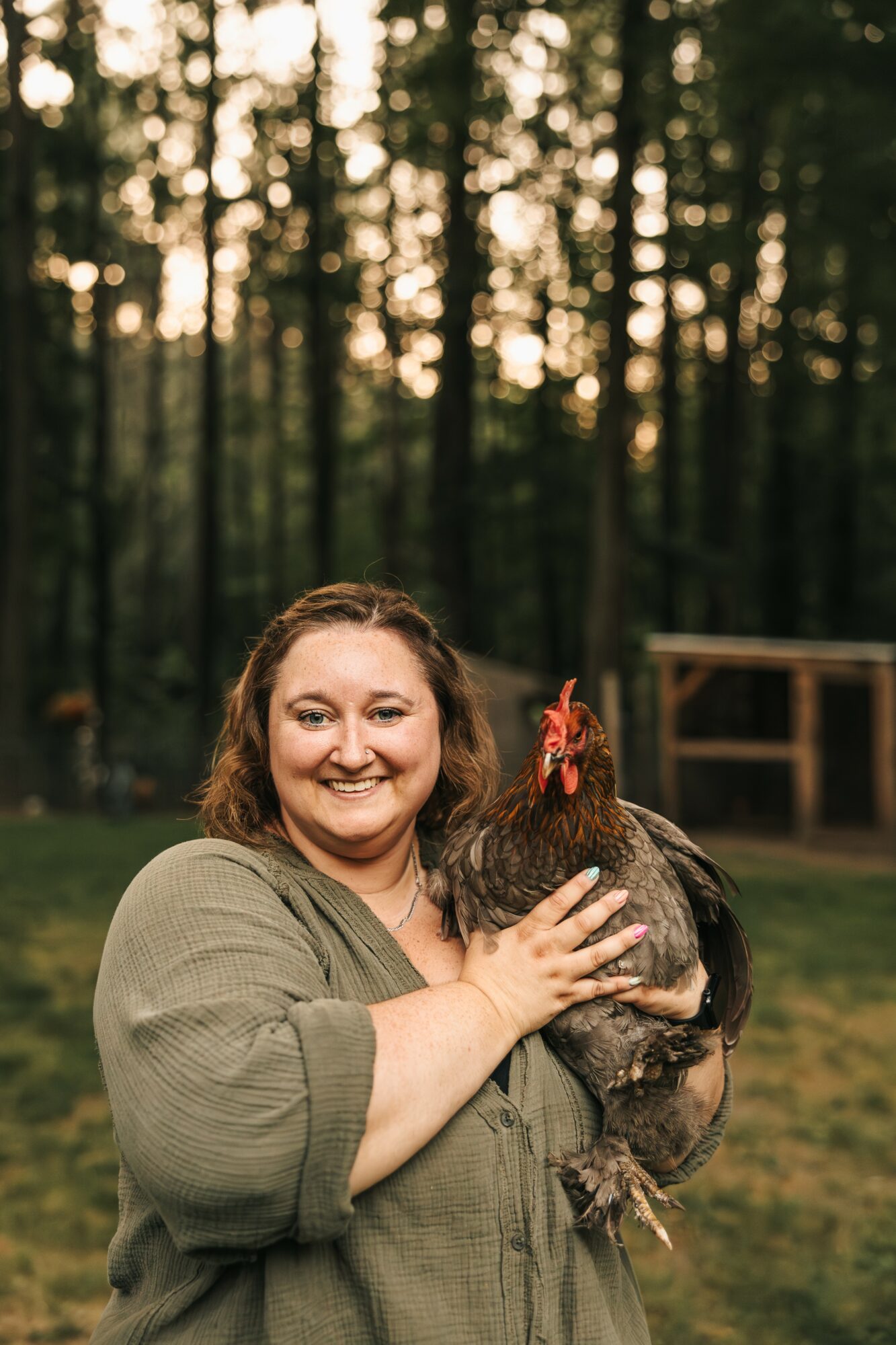 Woman smiling holding a chicken outdoors in a wooded area during sunset.