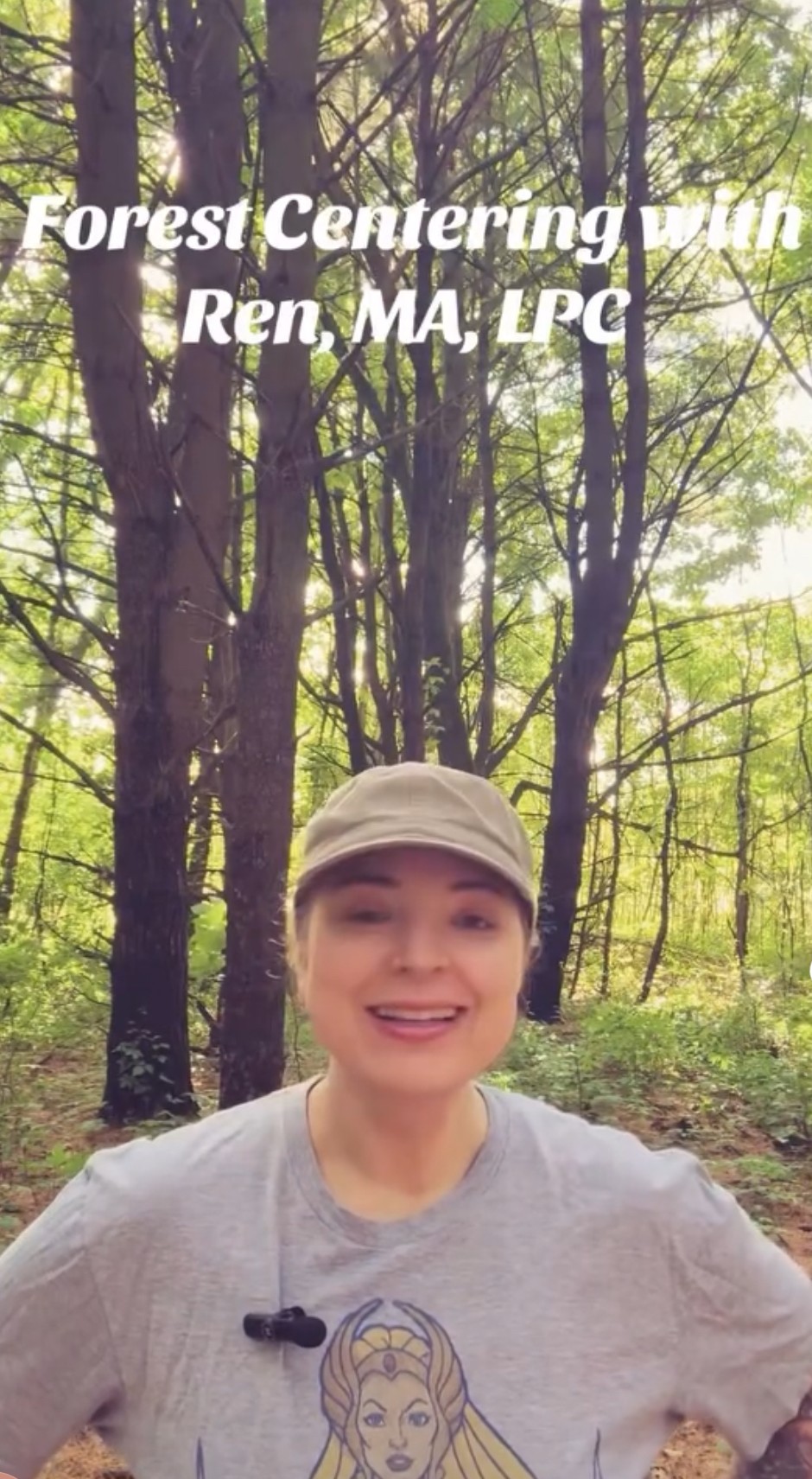 Person smiling outdoors in a forest, wearing a cap and a light-colored shirt with a graphic, trees in background.