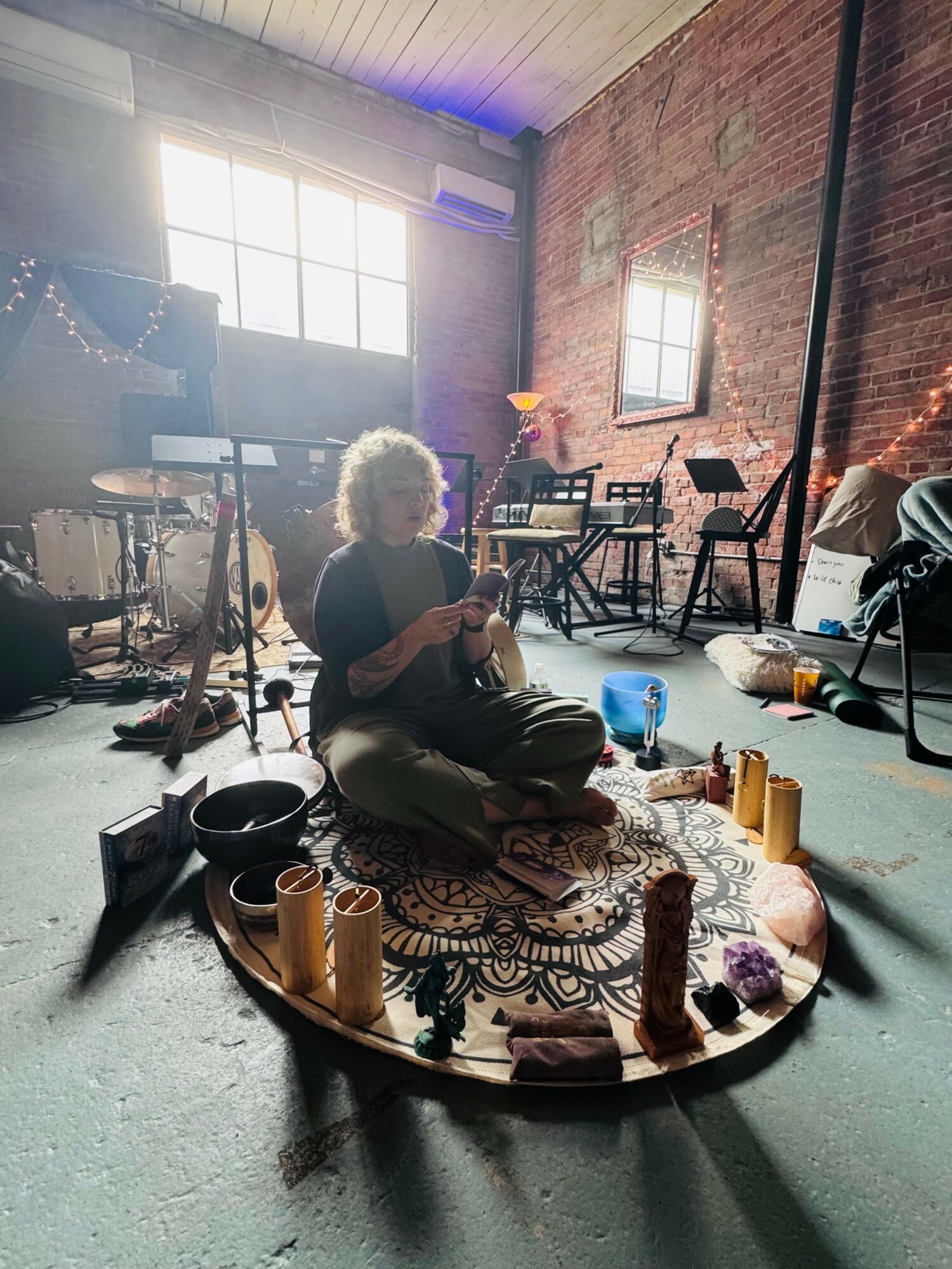 Person sitting cross-legged on a patterned rug in a room with brick and concrete walls, large window, and musical equipment.