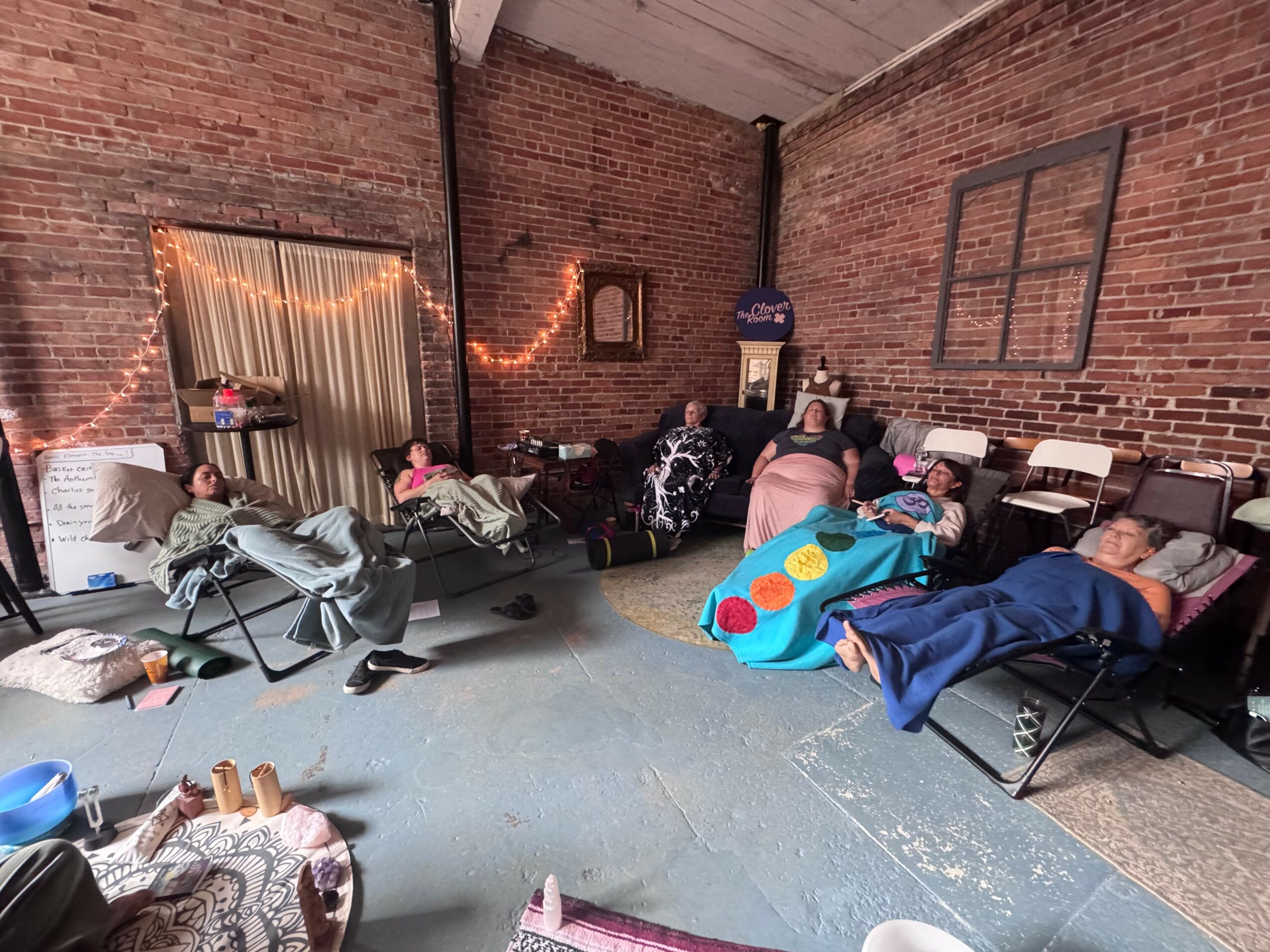 People sleeping on chairs and the floor in a brick-walled room with string lights and windows, cluttered with belongings.