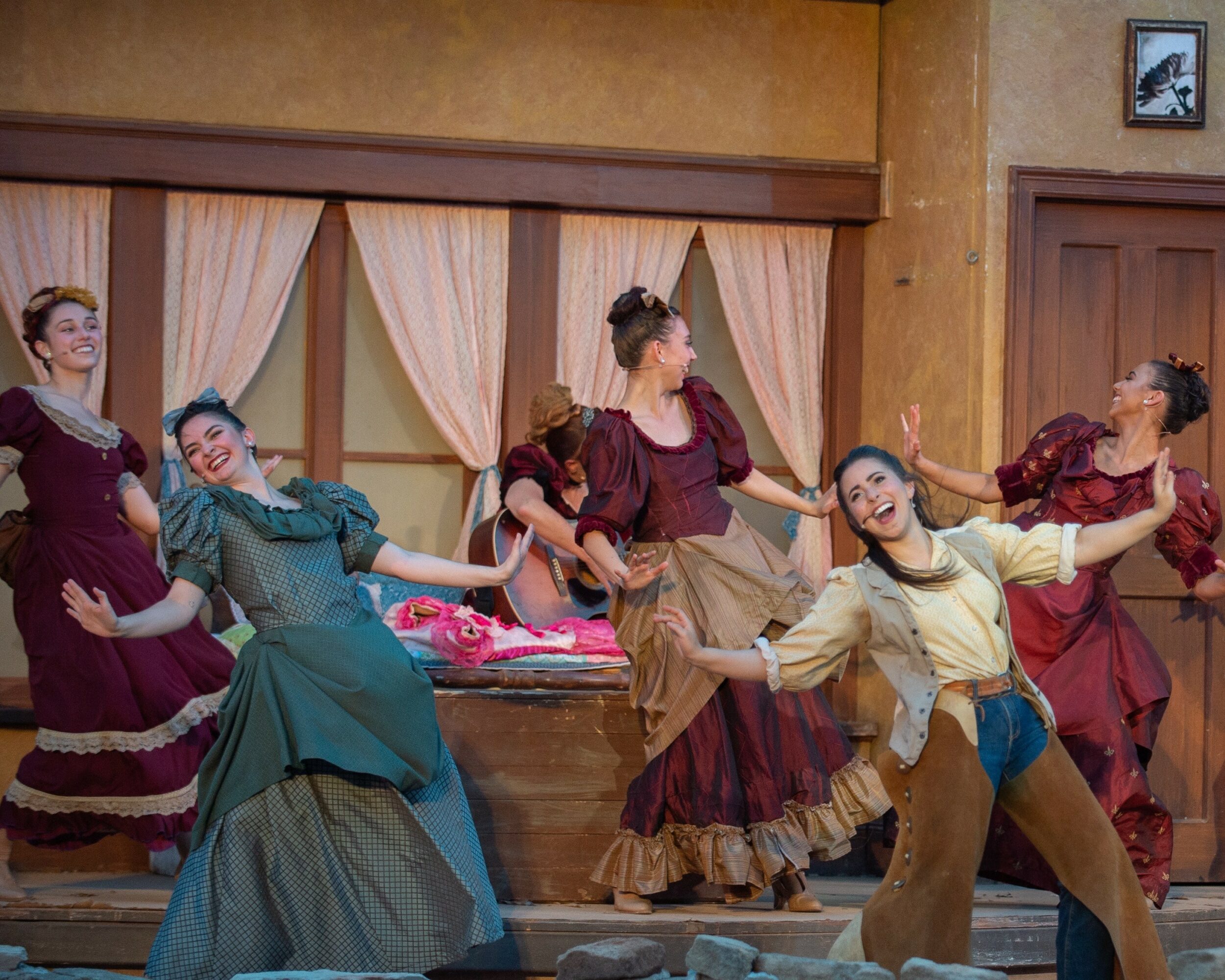 Group of women in vintage costumes dancing on stage with curtains in background.