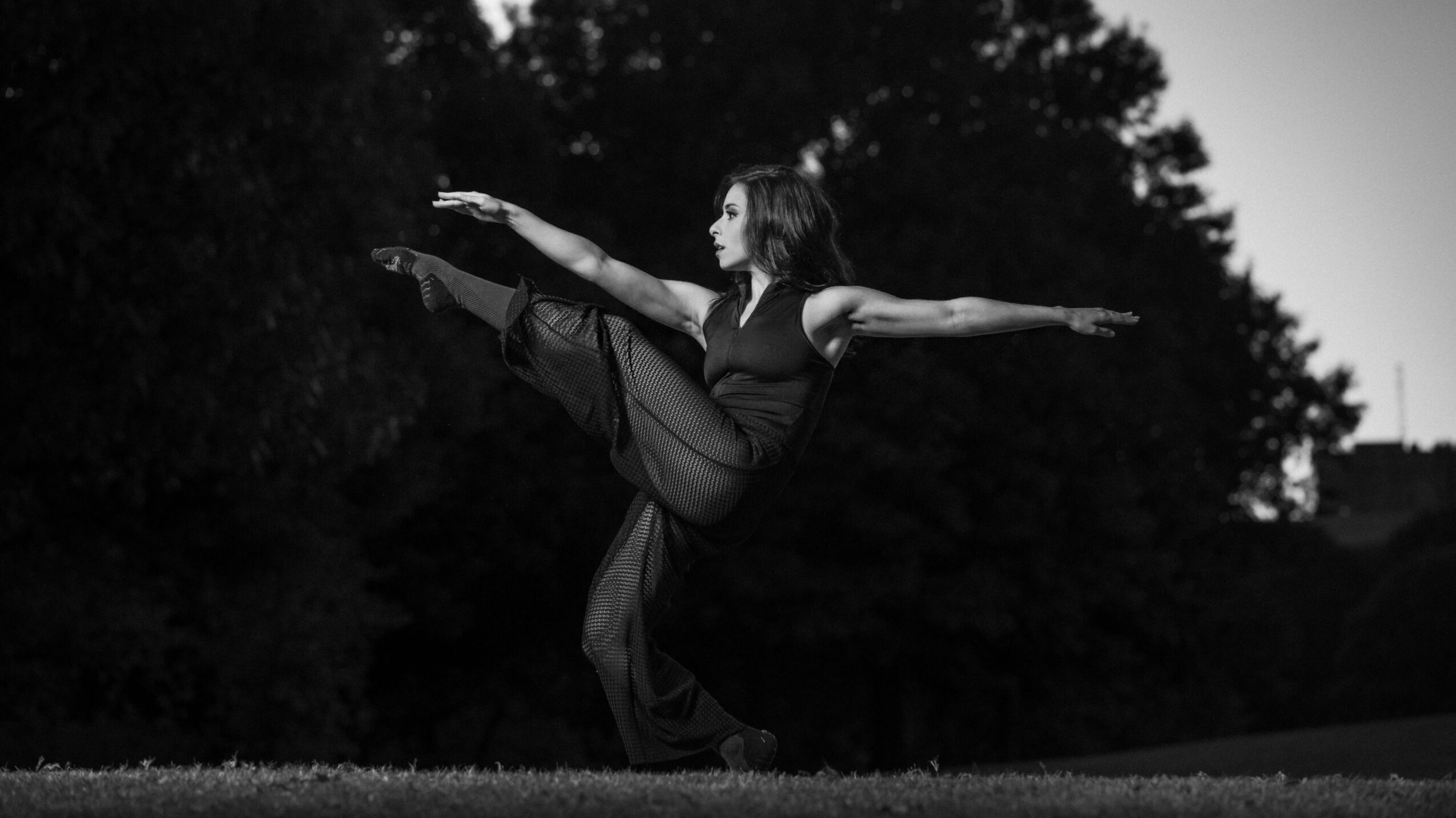 Person performing a high kick outdoors, arms extended, with trees in the background, in black and white.