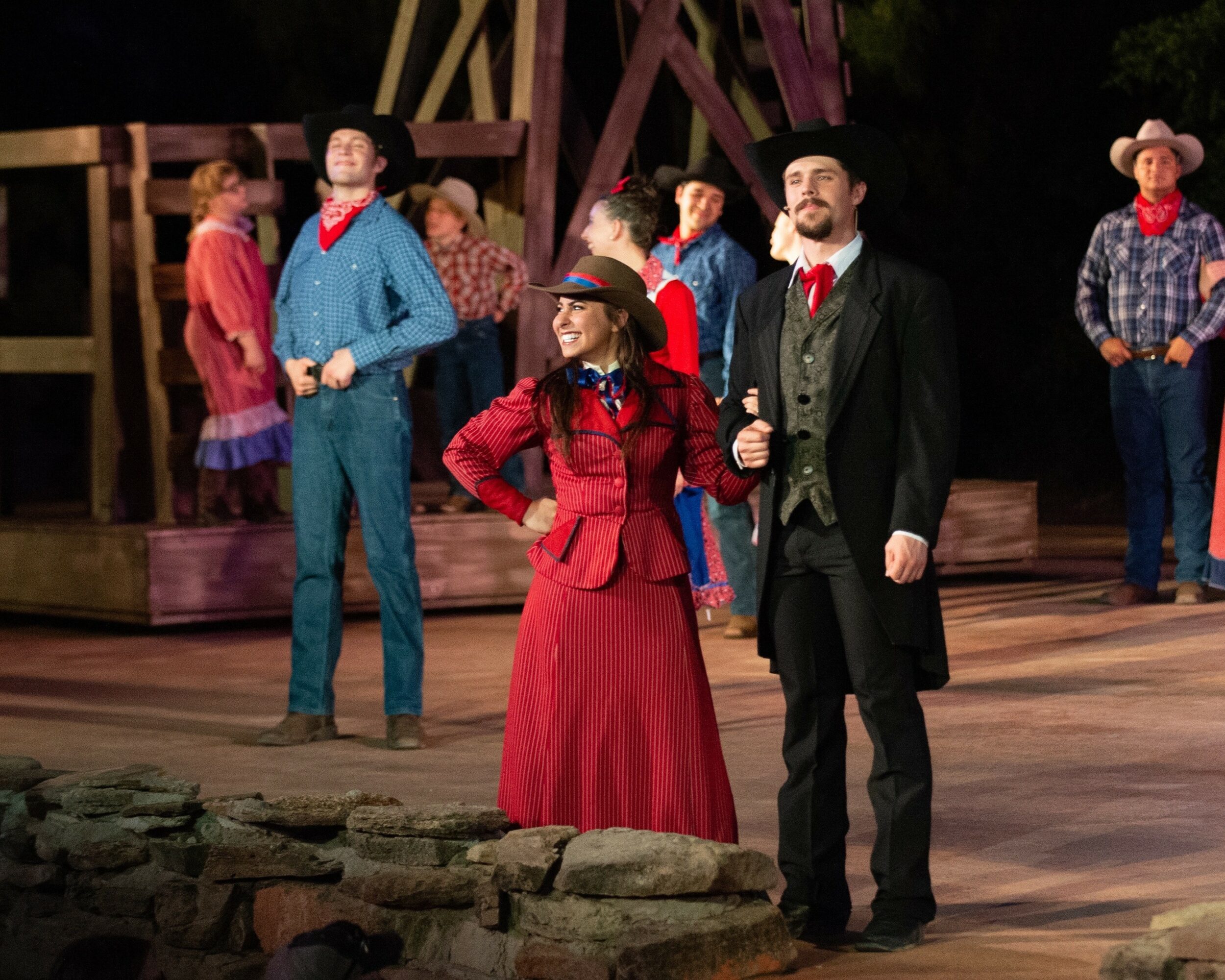 People in Western costumes on stage, some with hats, in a rural setting with wooden structures.