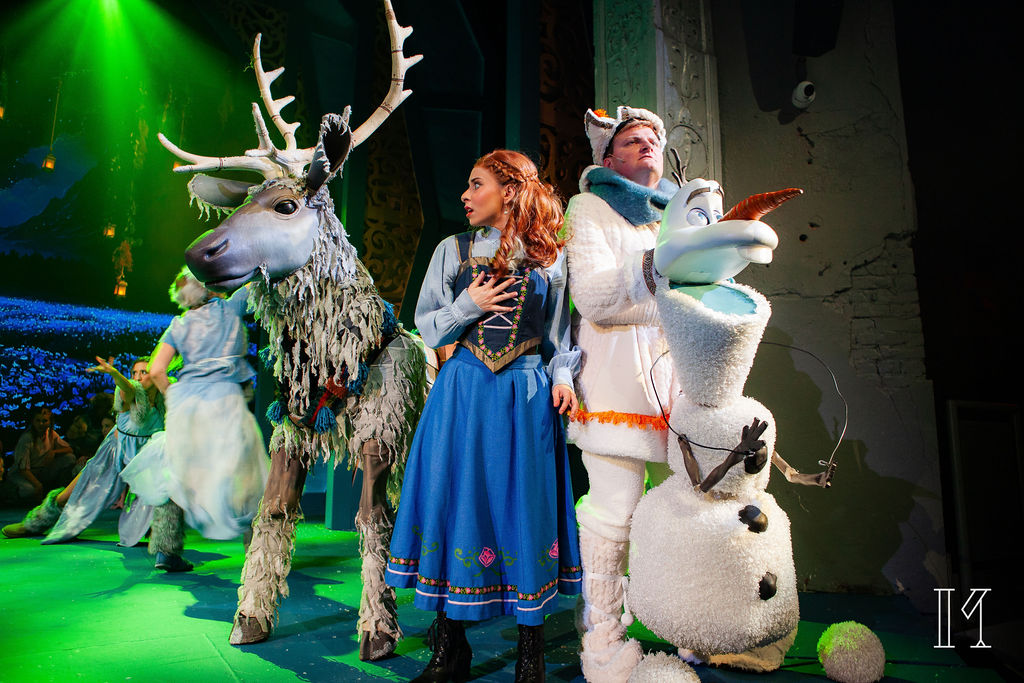 Three characters from a stage performance: a woman in a blue dress, a man in a white costume, and a snowman, with a reindeer in the background, on a green-lit stage.