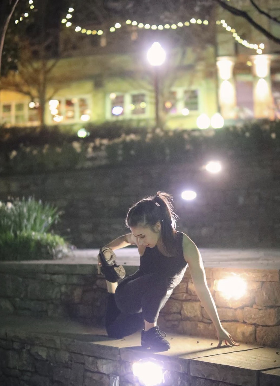 Young girl crouching on a stone ledge at night, illuminated by outdoor lights, with a building and string lights in background.