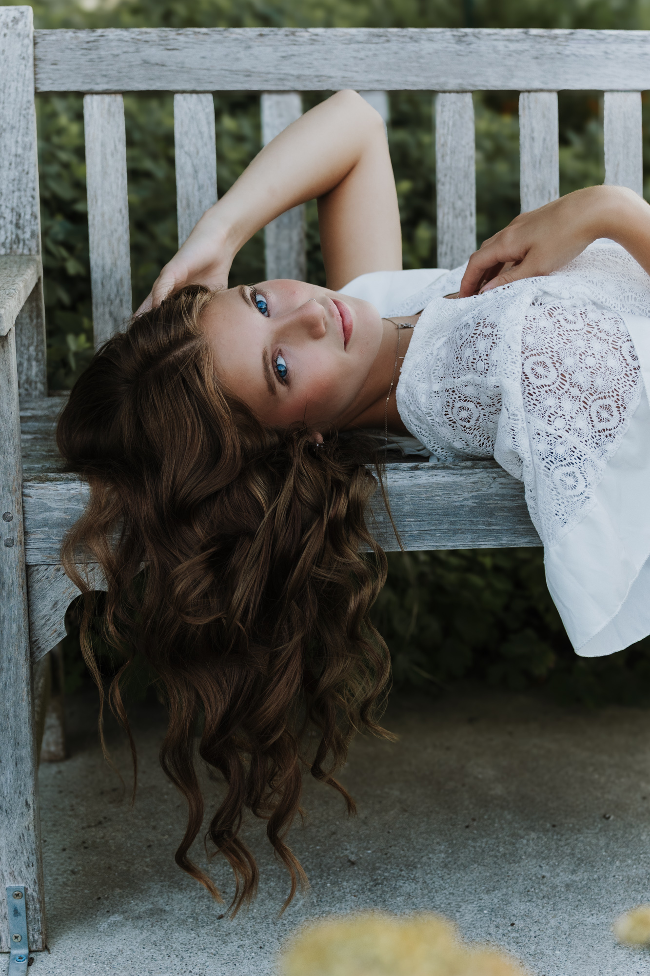 Woman with long, curly hair lying on a wooden bench outdoors, wearing a white lace dress, with her arm resting on her head.