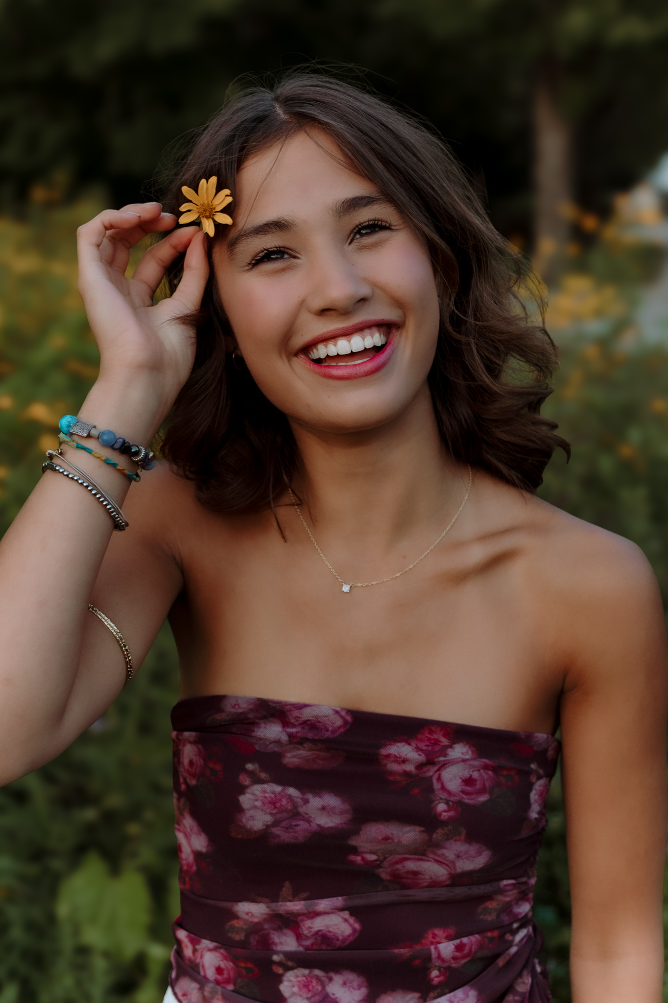 Smiling young woman holding a yellow flower near her head outdoors, wearing a strapless floral top, with trees in background.