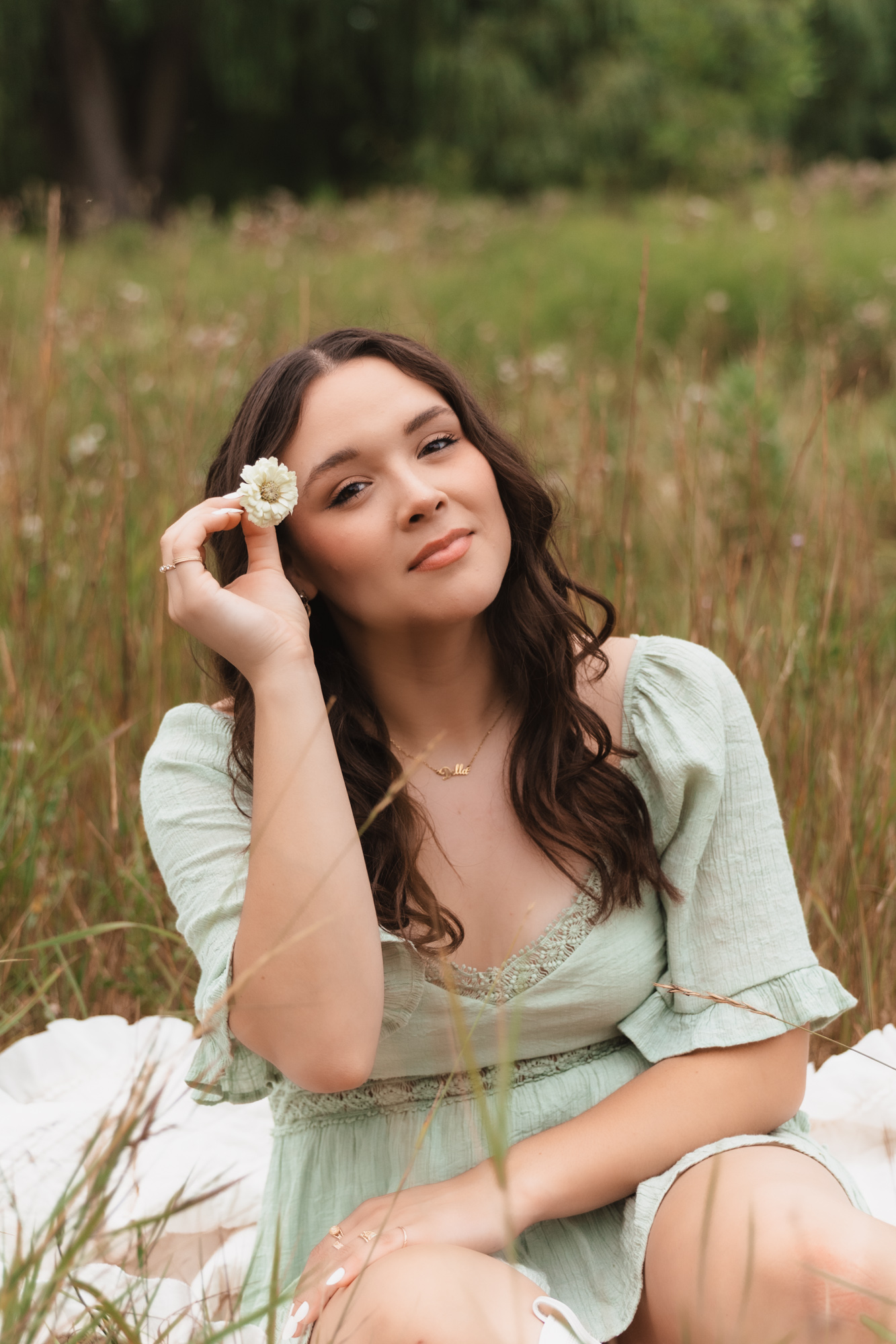 Young woman with long dark hair holding a flower near her face in a grassy field, smiling softly.