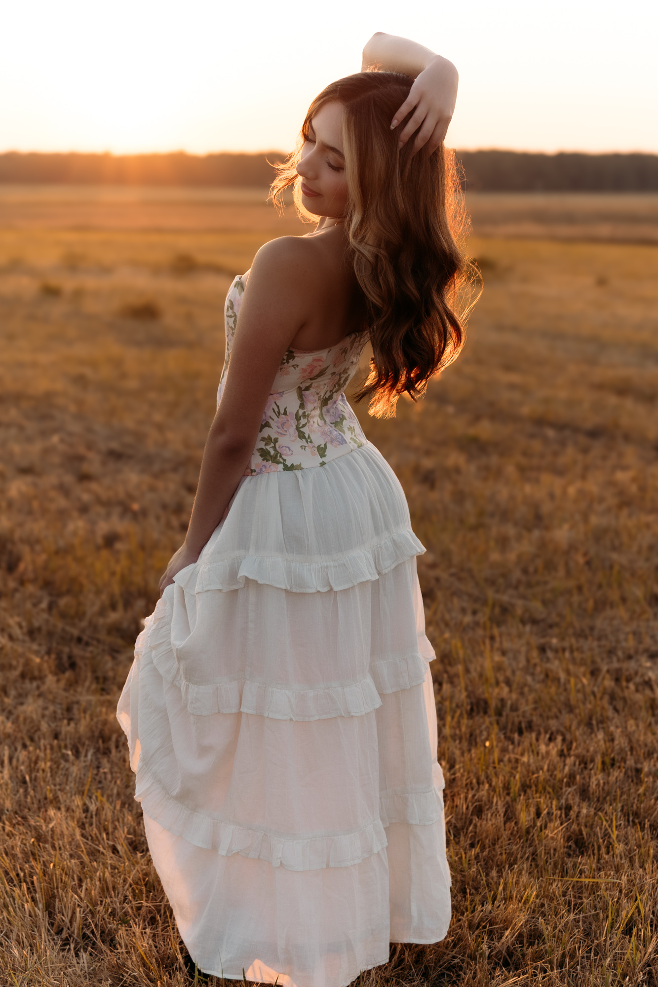Woman in a white dress standing in a field during sunset, with her hand on her head and looking to the side.