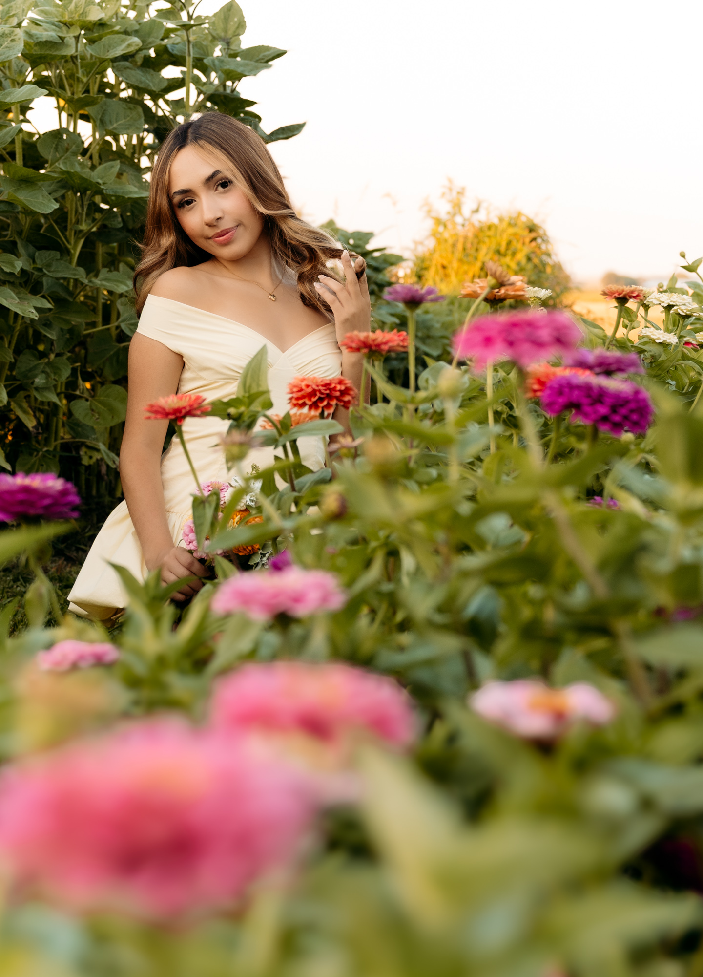 Young woman in a white dress standing among colorful flowers outdoors, smiling gently, with greenery and sky in background.