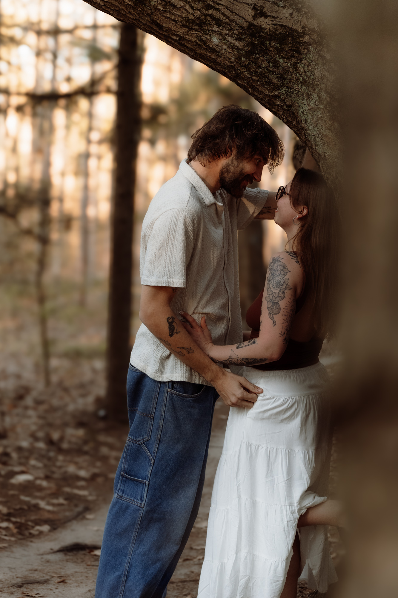 A couple stands close under a tree in a forest, gazing at each other affectionately, with sunlight filtering through trees.