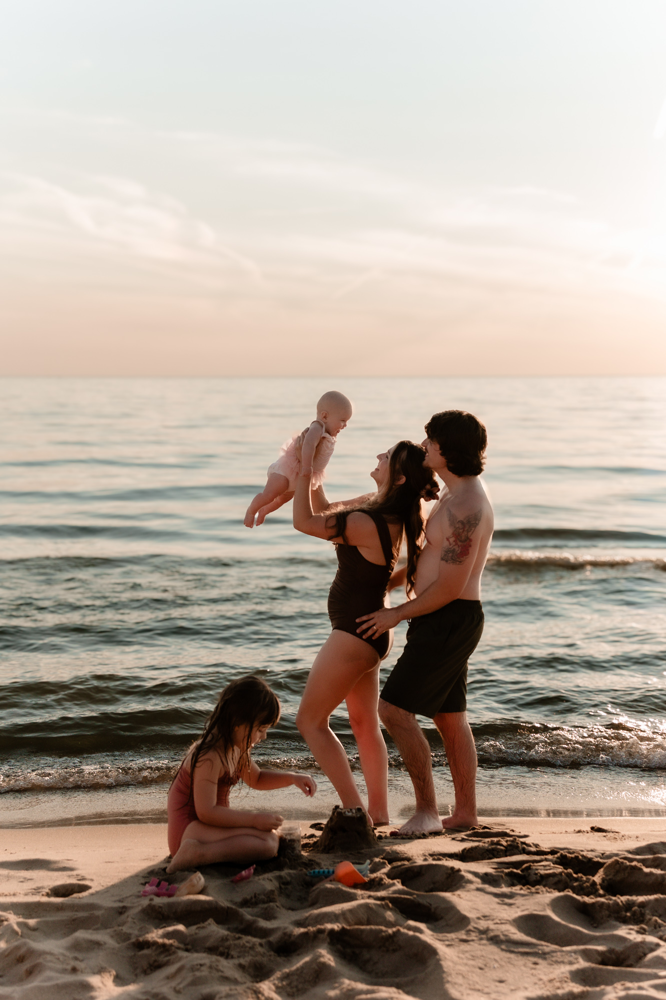 Four people, two adults and two children, on a beach near the water during sunset, with waves and sky visible.