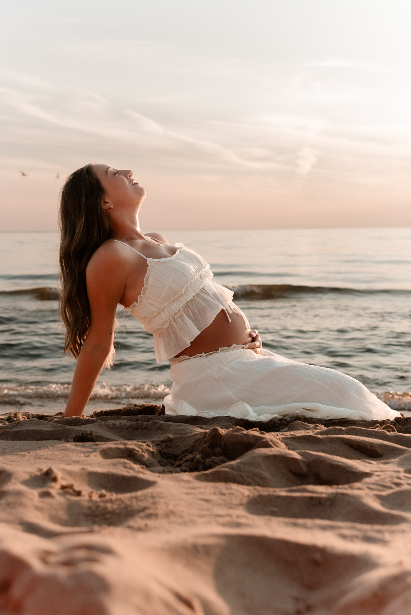 Pregnant woman in white dress sitting on beach sand, facing the ocean at sunset, with her head tilted back and eyes closed.