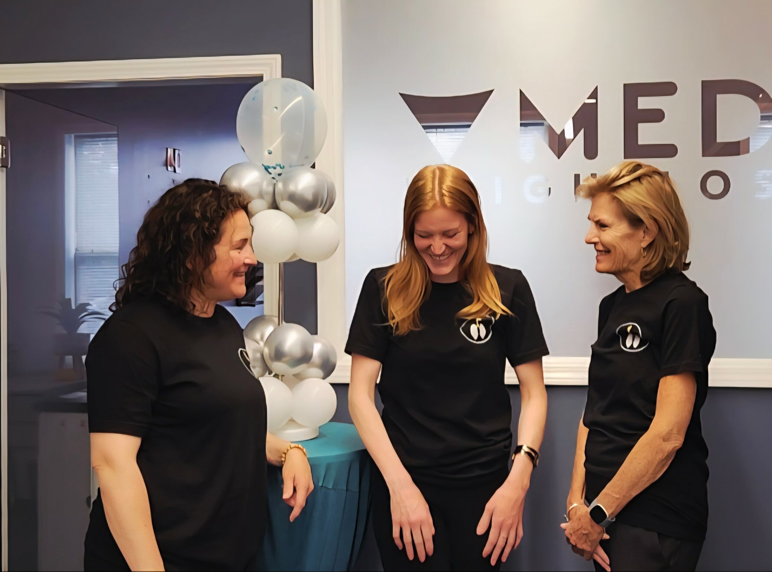 Three women in black shirts standing and talking indoors, with balloons and a logo in the background.