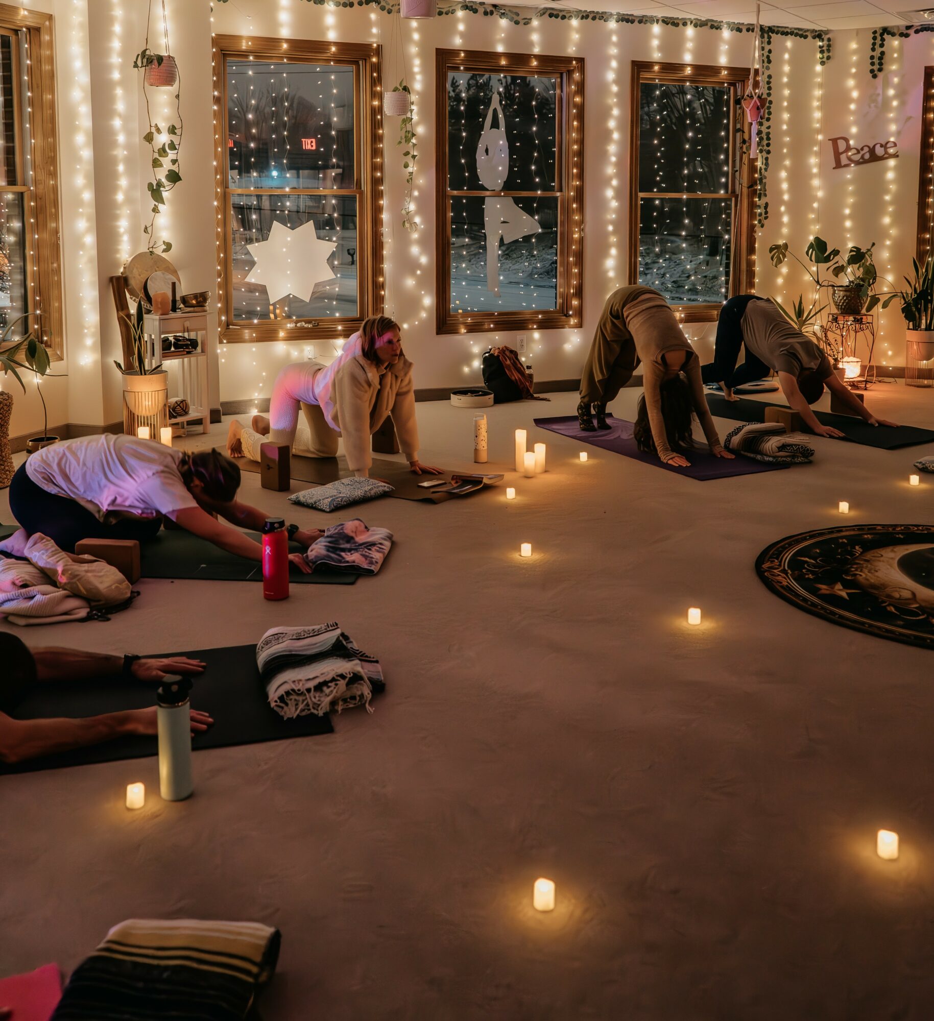 People practicing yoga in a warmly lit room with candles, decorated with string lights and large windows, during evening.