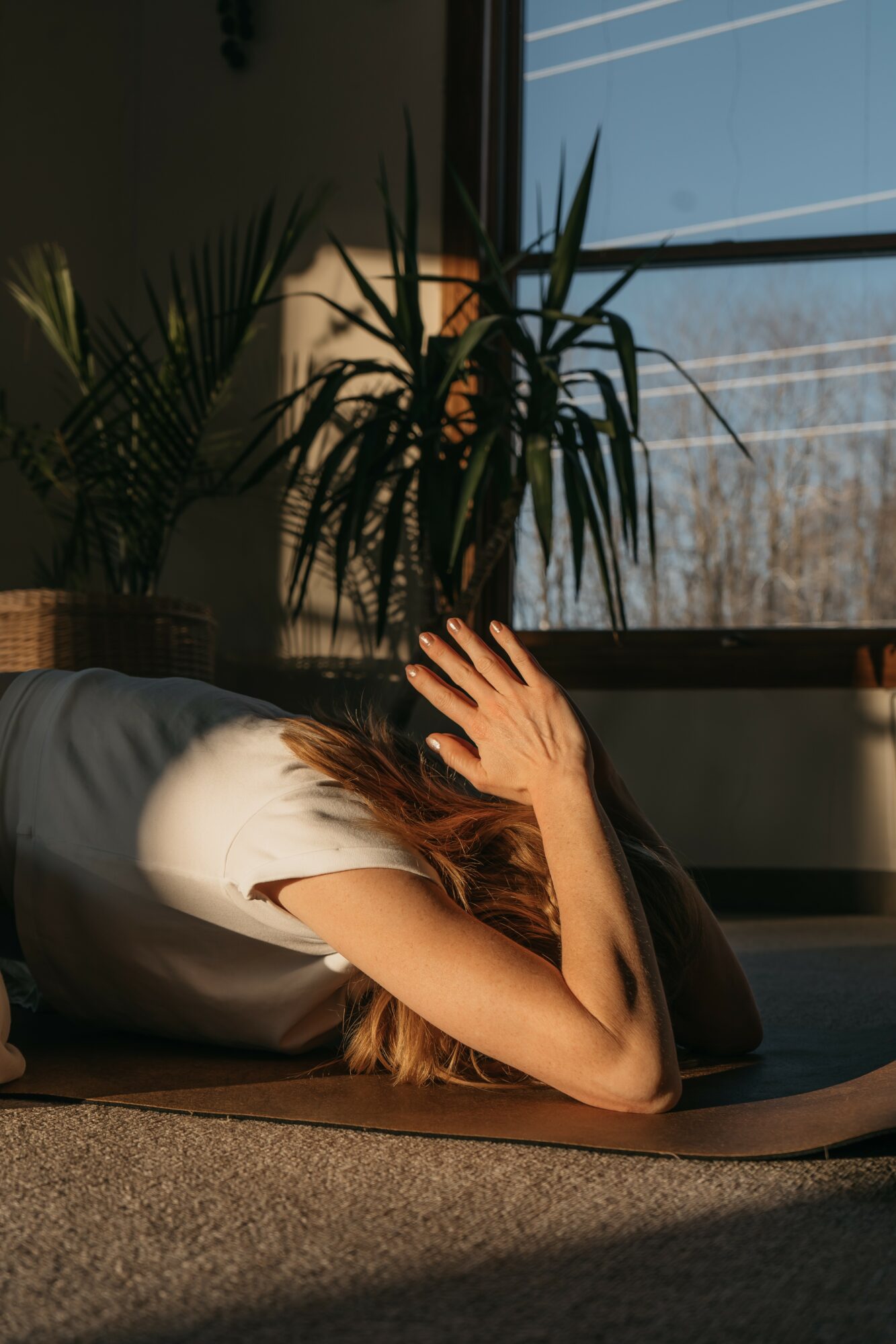 Person lying on the floor with hand covering face, sunlight streaming through window, plants in background.