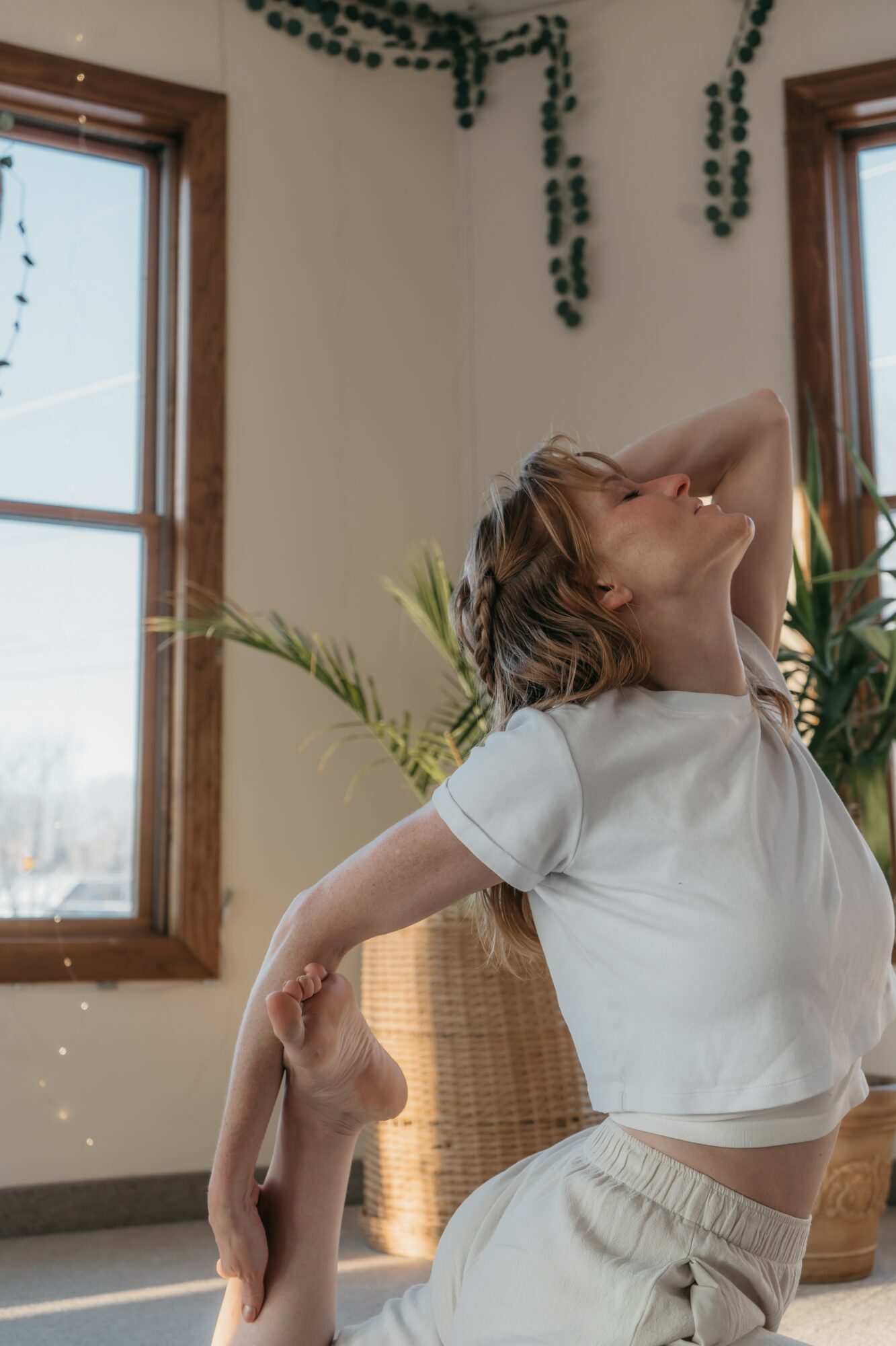 Woman stretching with her head tilted back and arm raised, in a room with large windows and plants.