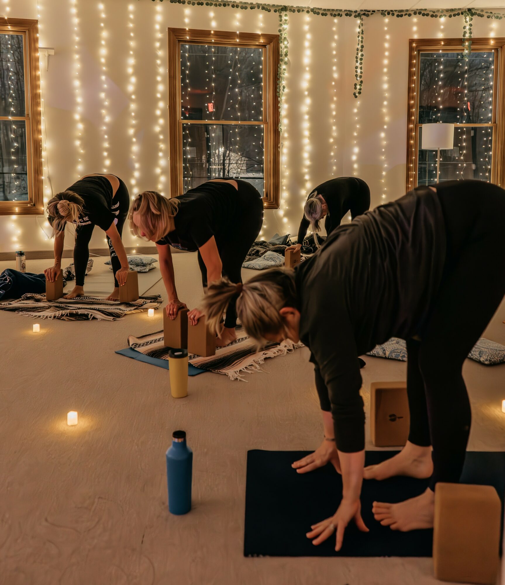 Four people practicing yoga or stretching on mats in a decorated room with string lights and windows.