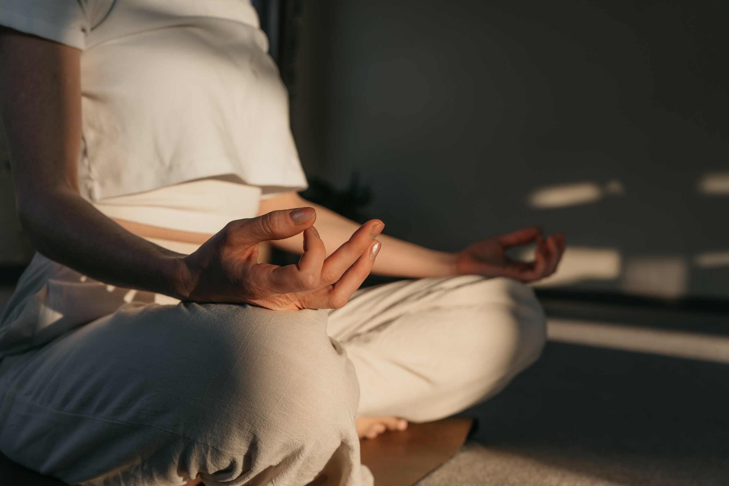 Person sitting cross-legged on the floor, practicing meditation with hand in a mudra gesture, in a dimly lit room.