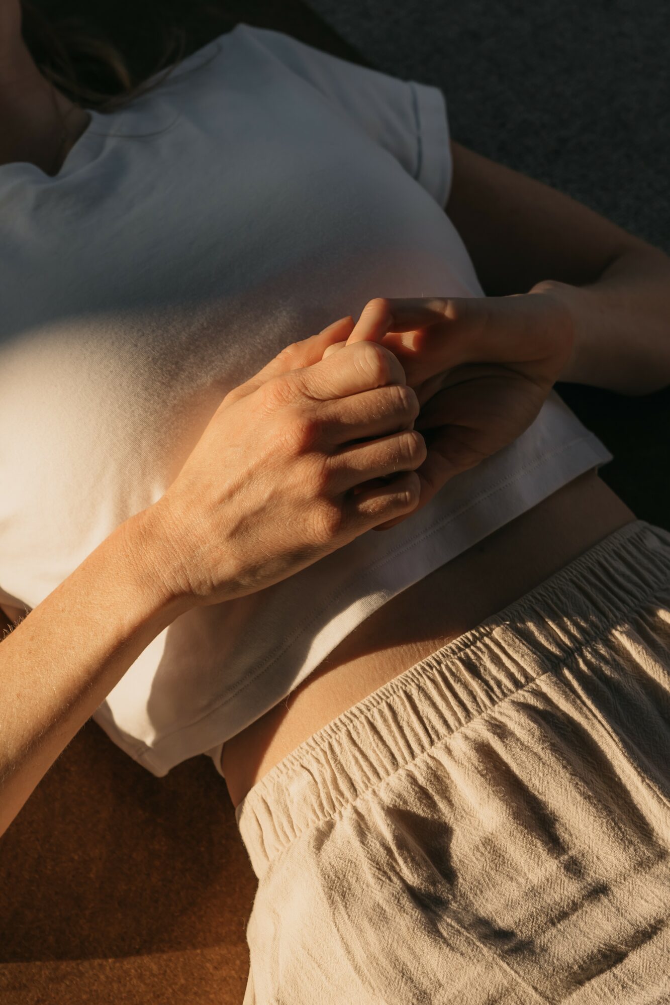 Person lying down, holding hands with another person, wearing a white shirt and beige pants, sunlight on skin.