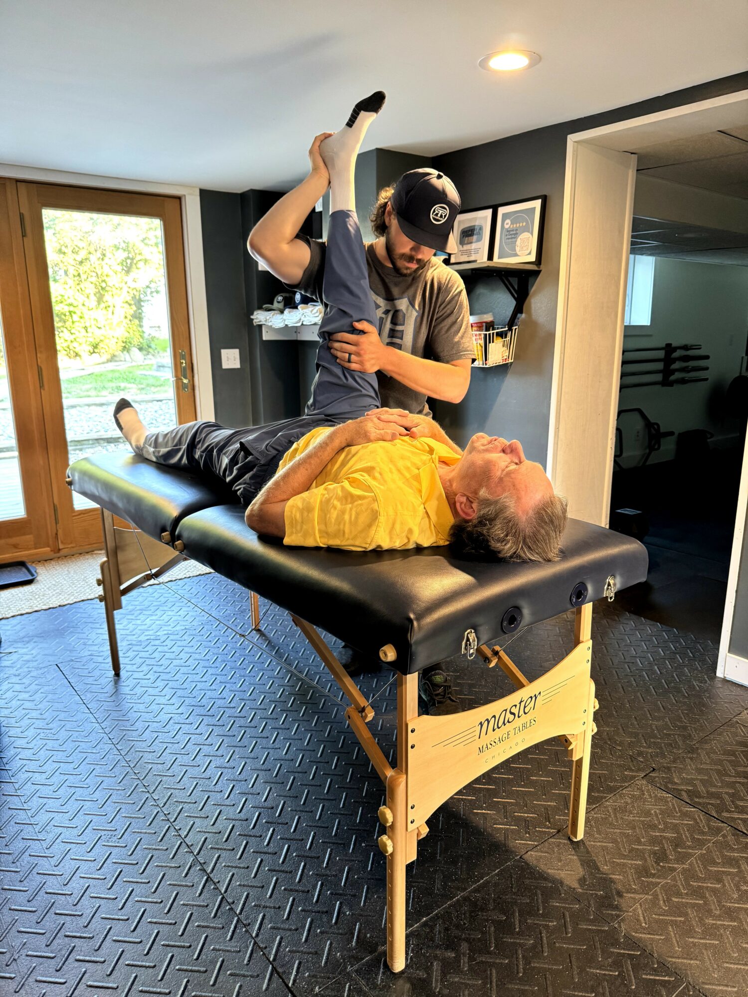 A person lying face up on a massage table with a yellow shirt, receiving a treatment from a practitioner in a room with a window and framed pictures.